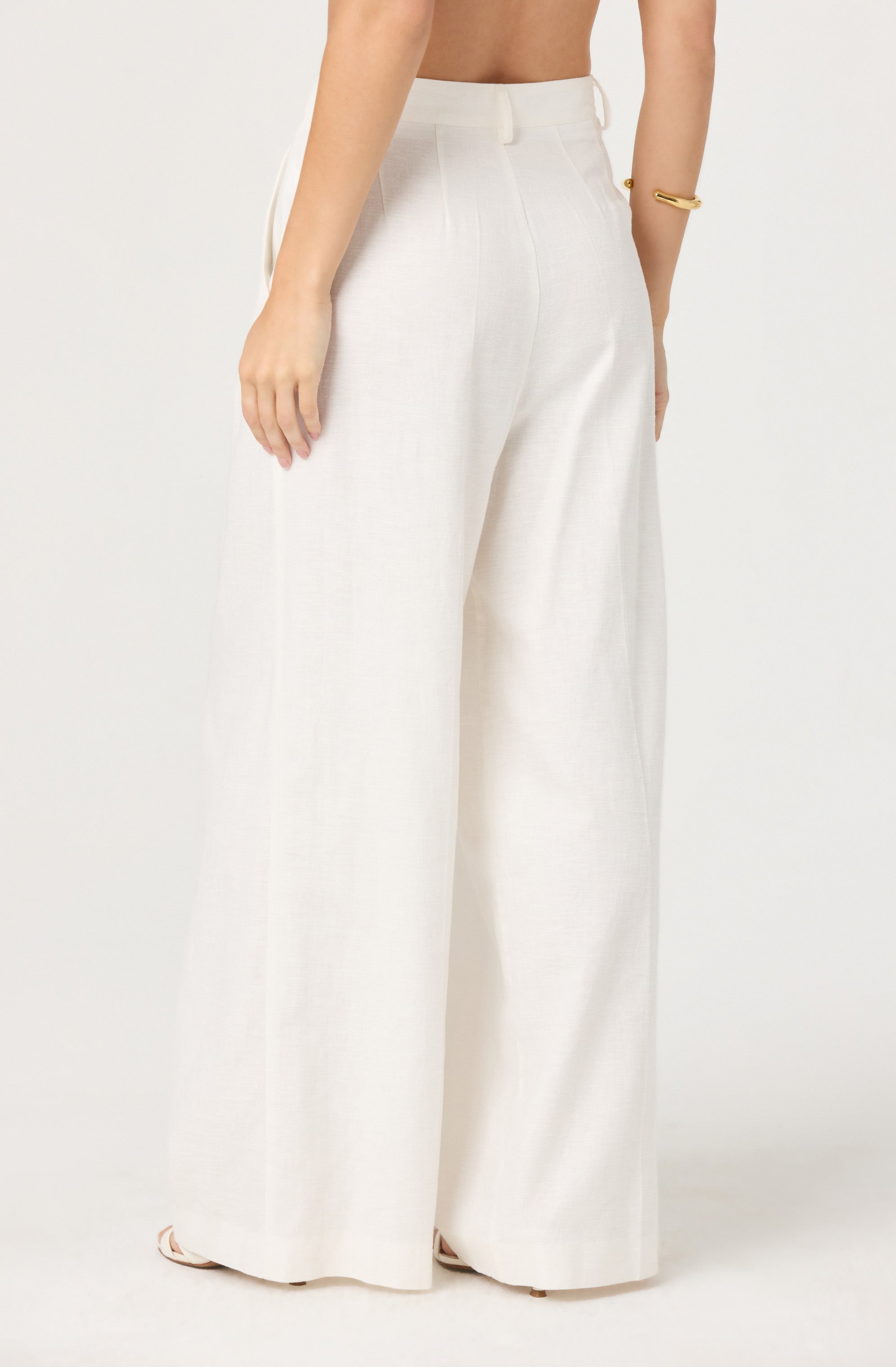 A person is wearing high-waisted, wide-leg white trousers made of a light, textured fabric. The trousers have a clean waistband with no visible belt, and they extend down to cover the shoes. The person is accessorizing with a simple gold bracelet on their right wrist.