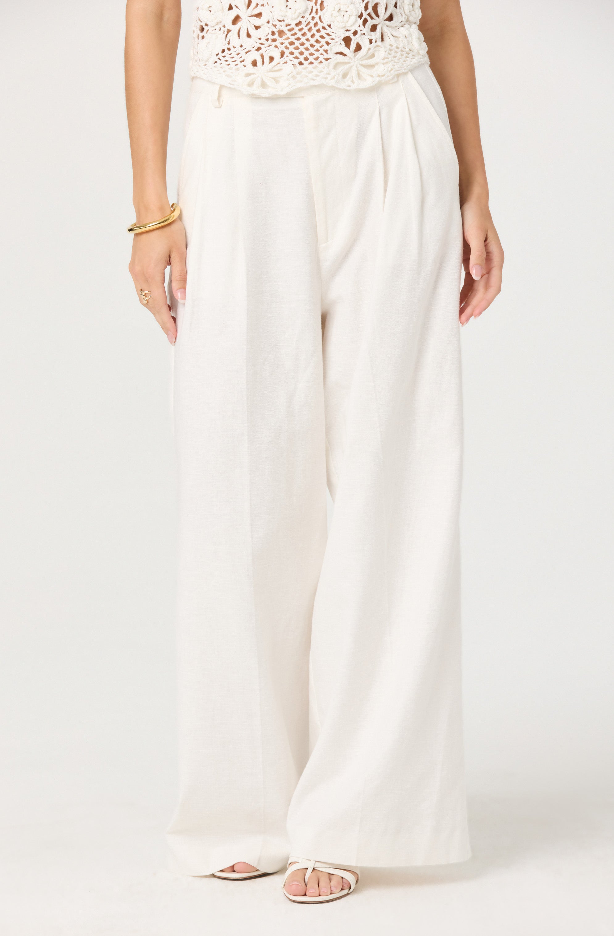 A person is wearing high-waisted, wide-leg white trousers made of a lightweight, flowing fabric. The trousers have a pleated front and are paired with a crochet top that features a floral pattern. The person accessorizes with gold bangles on their right wrist, a ring, and open-toe sandals.