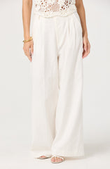 A person is wearing high-waisted, wide-leg white trousers made of a lightweight, flowing fabric. The trousers have a pleated front and are paired with a crochet top that features a floral pattern. The person accessorizes with gold bangles on their right wrist, a ring, and open-toe sandals.