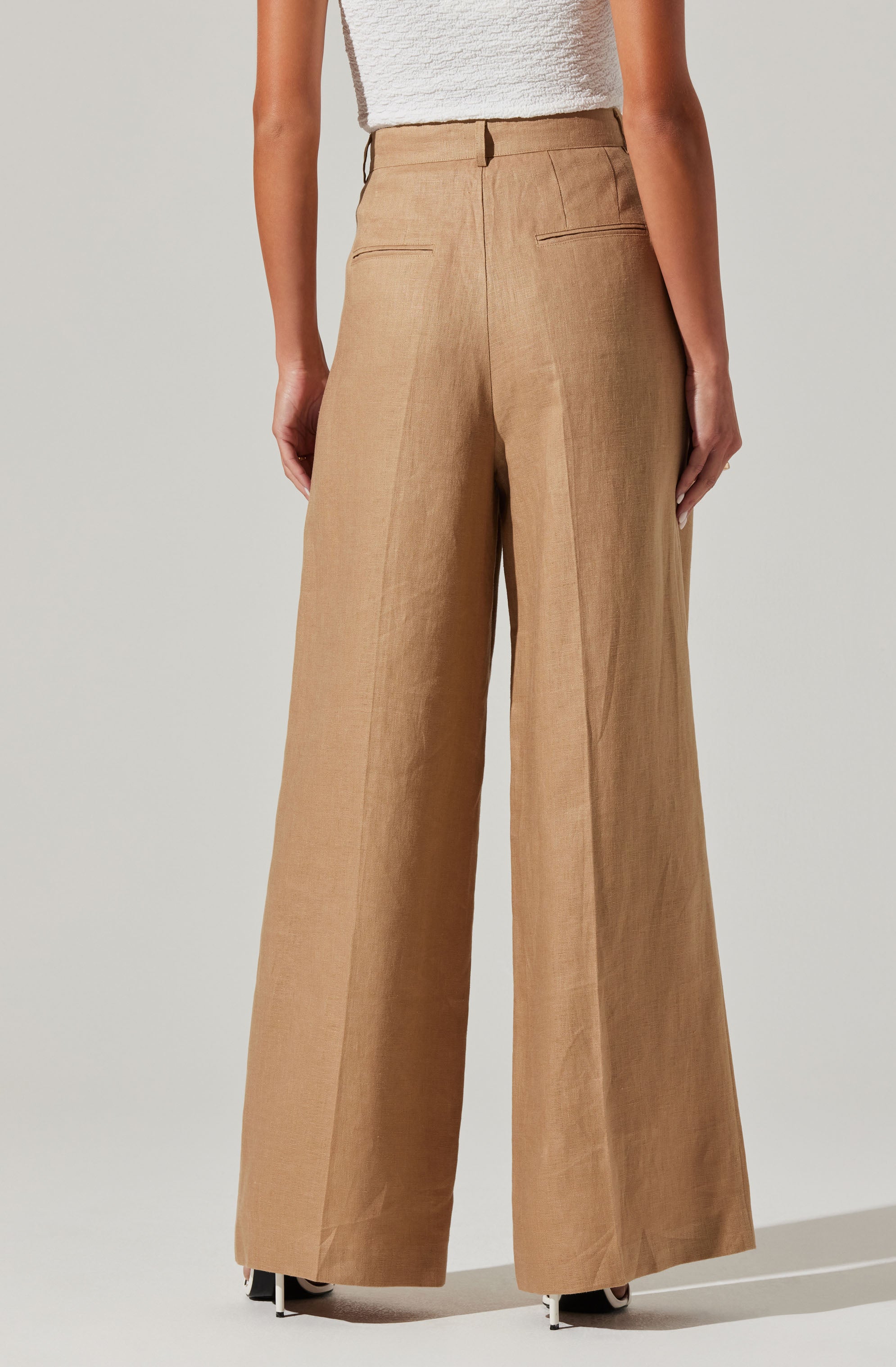 The image shows a person wearing beige, high-waisted, wide-leg trousers with back welt pockets, paired with a white textured sleeveless top.