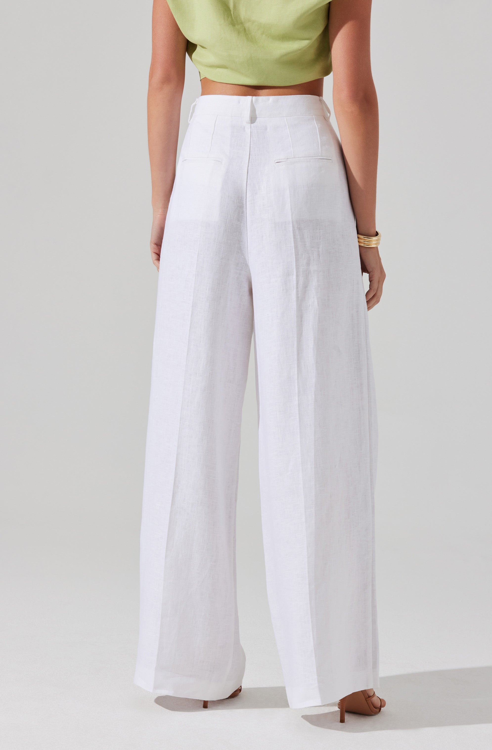 A person is wearing high-waisted, wide-leg white linen pants with visible front pockets, paired with a light green sleeveless crop top. They are also wearing tan high-heeled sandals and a stack of gold bracelets on their right wrist.