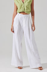 A person is wearing a sleeveless, light green, cropped blouse with a collar and a front knot. They are also wearing high-waisted, wide-leg white linen pants with pleats at the waist. The outfit is accessorized with multiple gold rings on their fingers and a gold bracelet on their wrist.