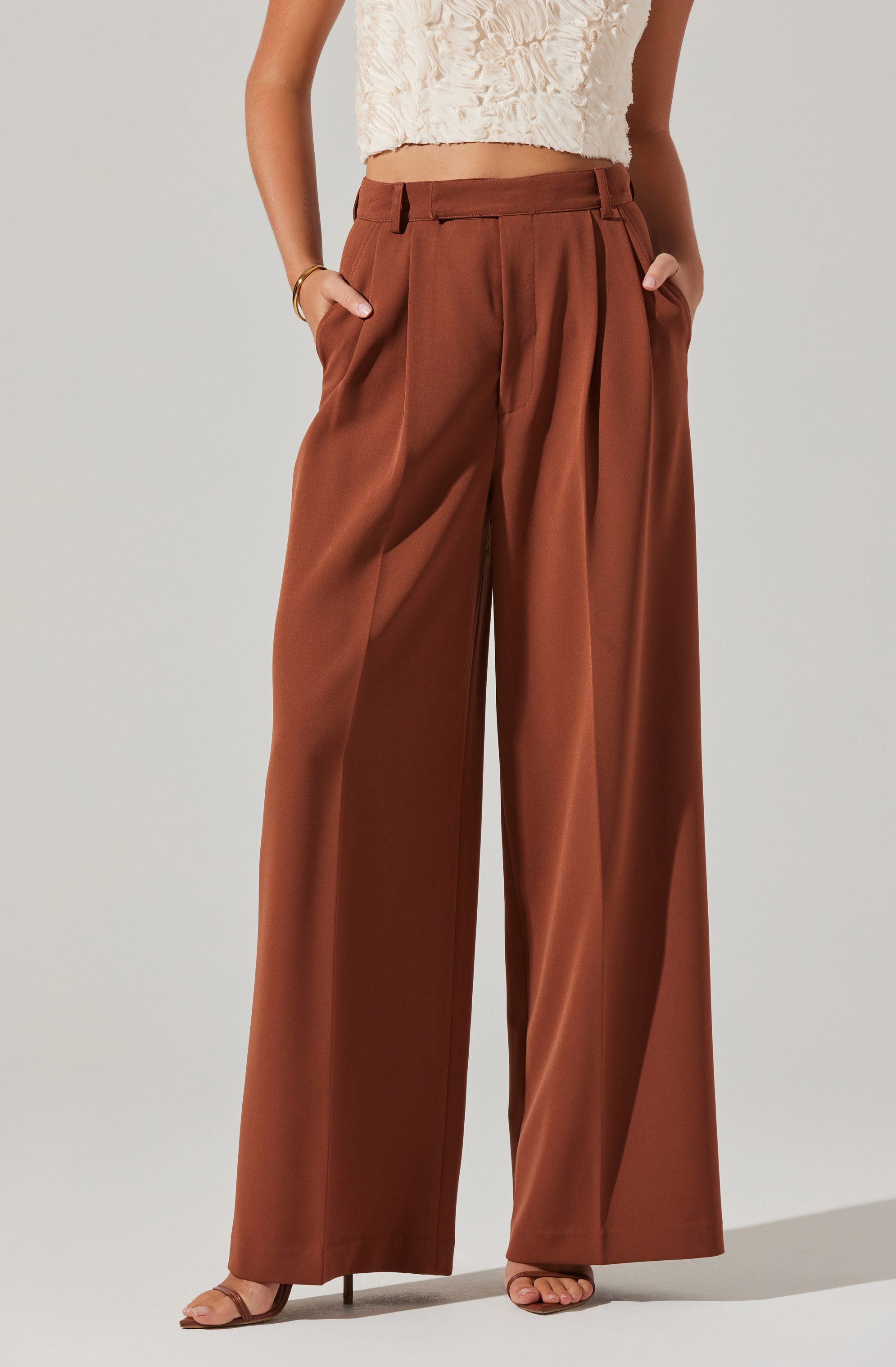 A person is wearing high-waisted, wide-leg brown trousers with pleats at the front and side pockets. The trousers have belt loops, but no belt is present. The individual is also wearing a cream-colored, textured sleeveless top with a floral or ruffled pattern. They have a gold bracelet on their left wrist and are wearing beige open-toed shoes.