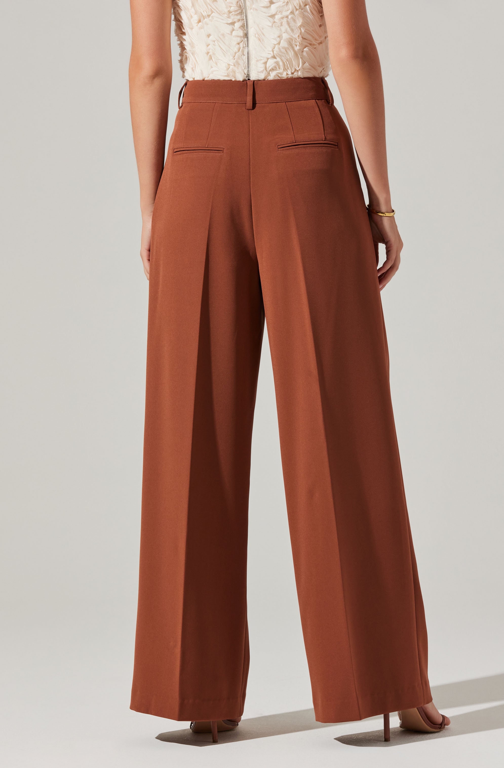 The image shows a person wearing high-waisted, wide-leg brown trousers with a smooth, tailored fit. The trousers have belt loops at the waistband and two horizontal welt pockets on the back. The person is also wearing a sleeveless top with textured fabric in a light color and a gold bracelet on their right wrist.