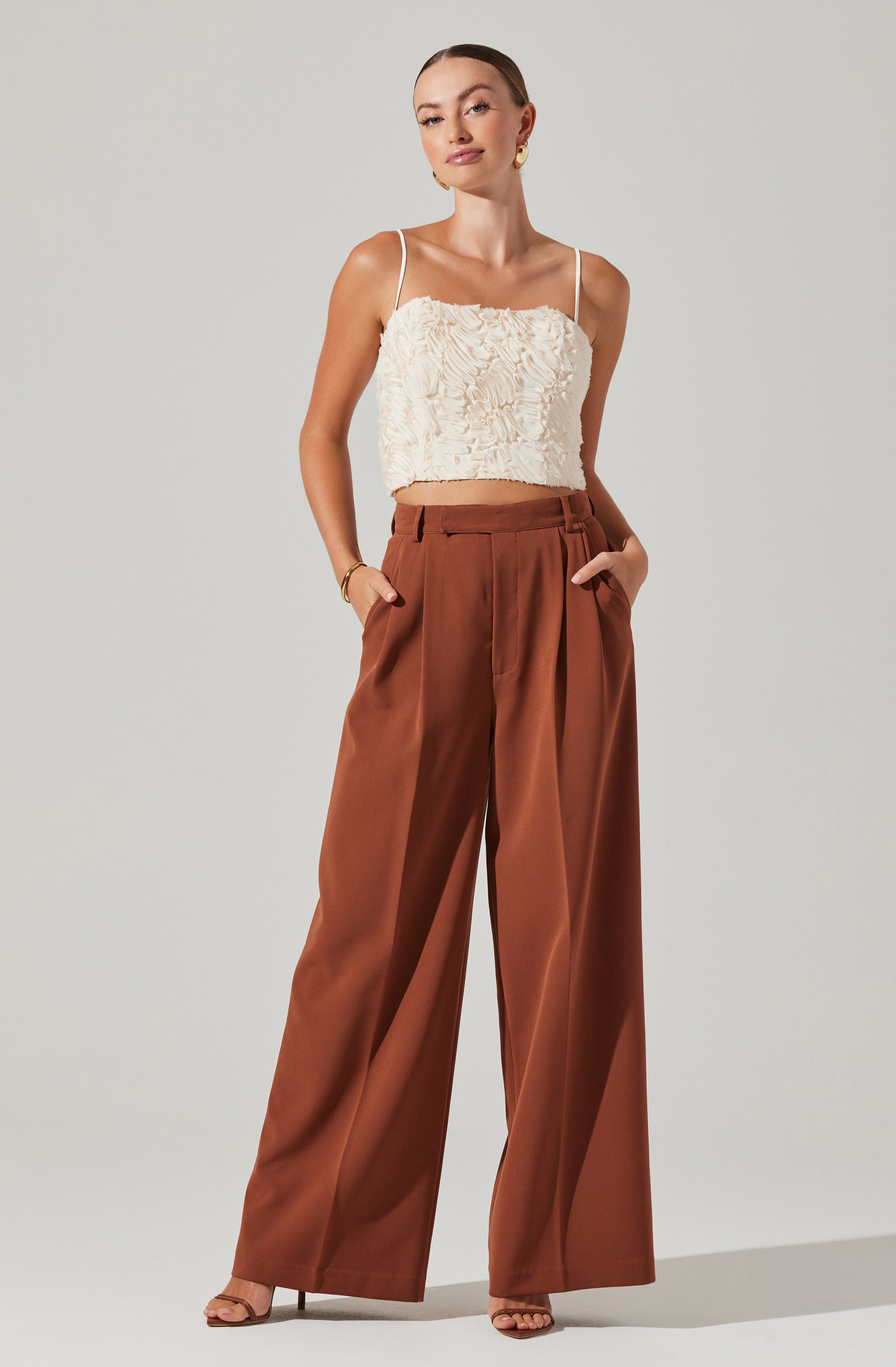 The woman is wearing a cream-colored textured crop top with thin spaghetti straps, paired with high-waisted, wide-leg brown trousers.