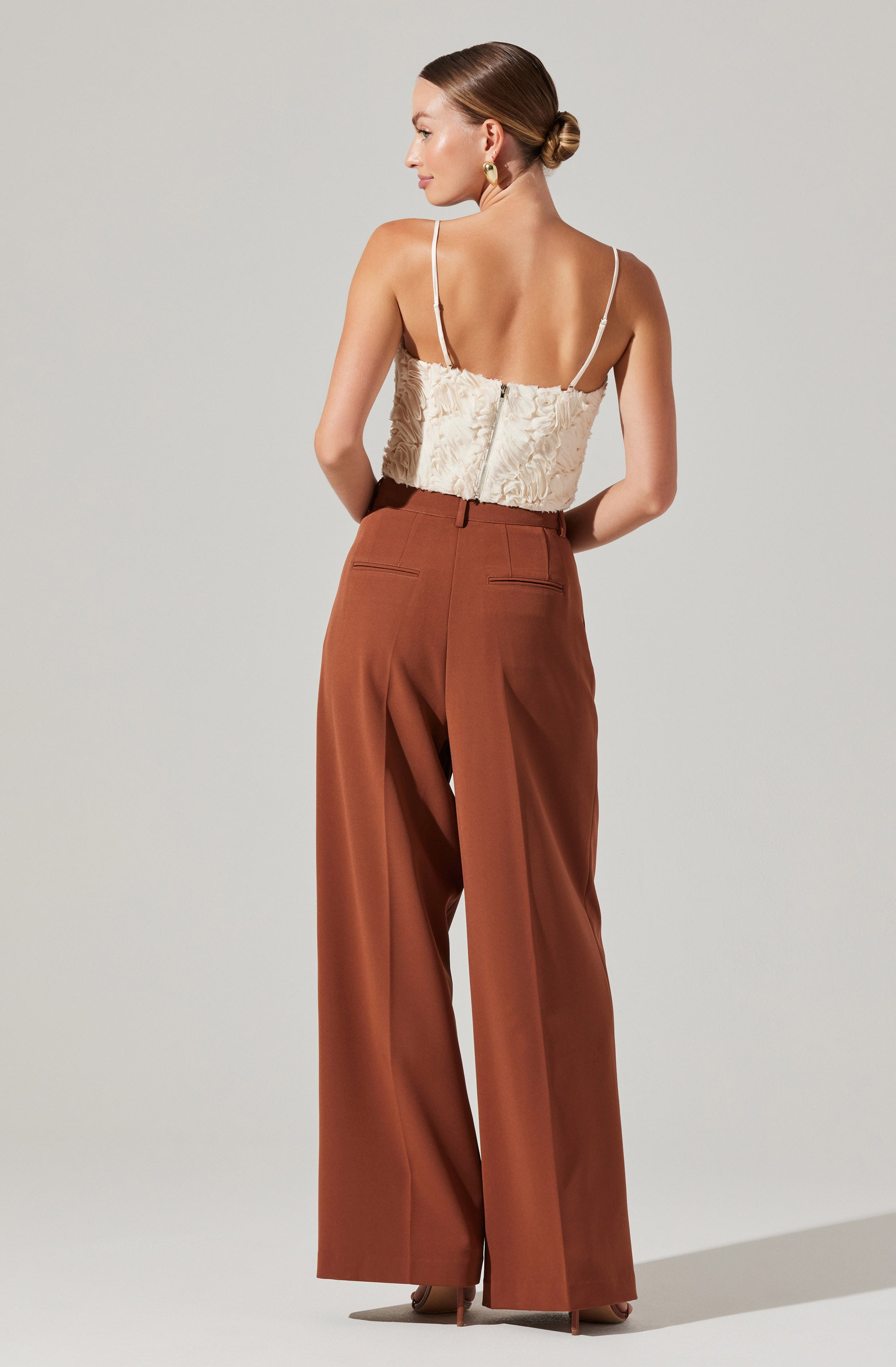 The woman is wearing a cream-colored, textured, strapless top with thin shoulder straps and a zipper at the back. She pairs it with high-waisted, wide-leg brown trousers that have a tailored fit and back welt pockets.