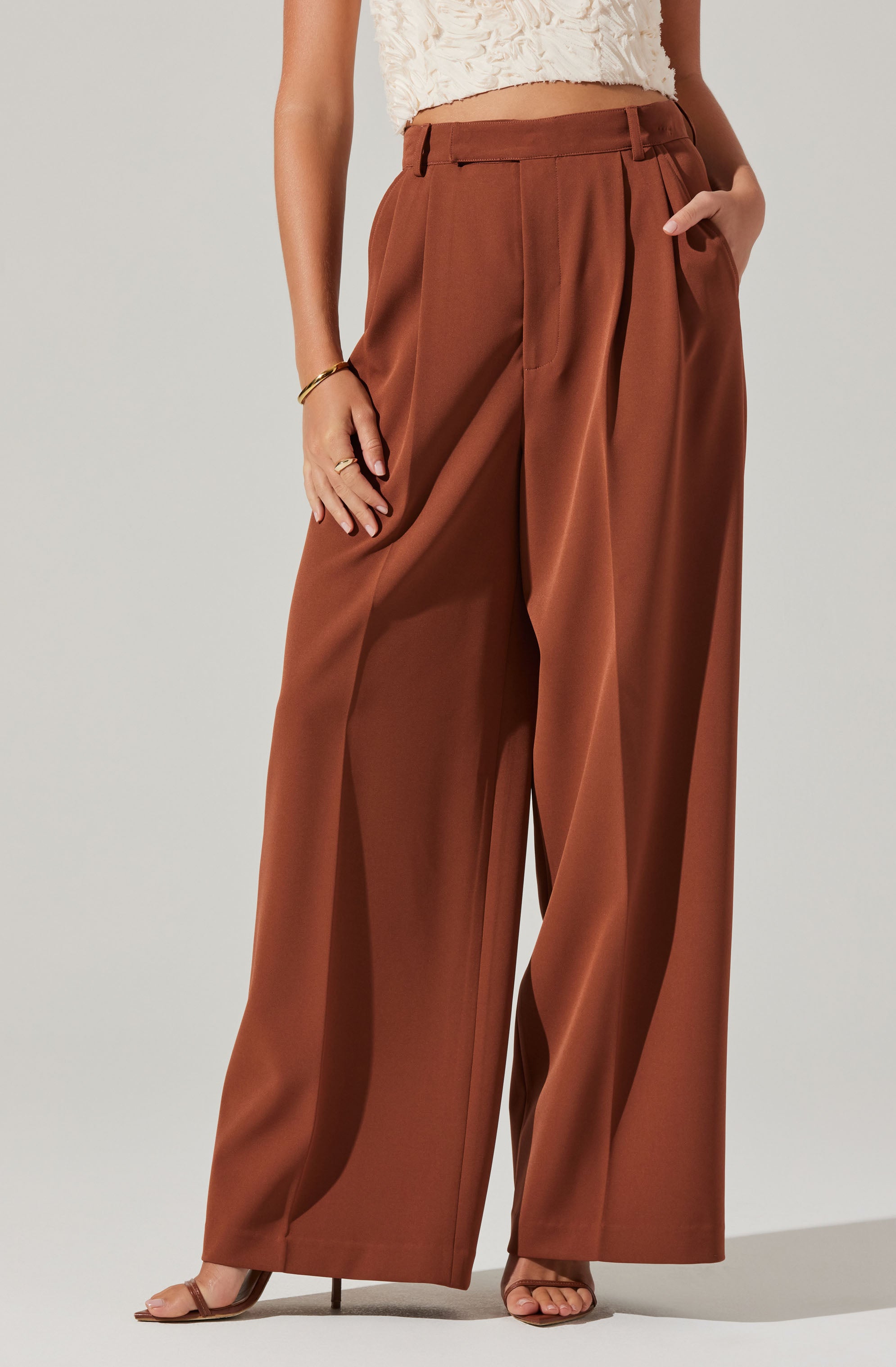 A person is wearing high-waisted, wide-leg trousers in a warm brown color, with pleats at the front. The trousers feature belt loops and side pockets. They are paired with a sleeveless, textured white top and open-toed heeled sandals. The person also has a gold bracelet and rings.