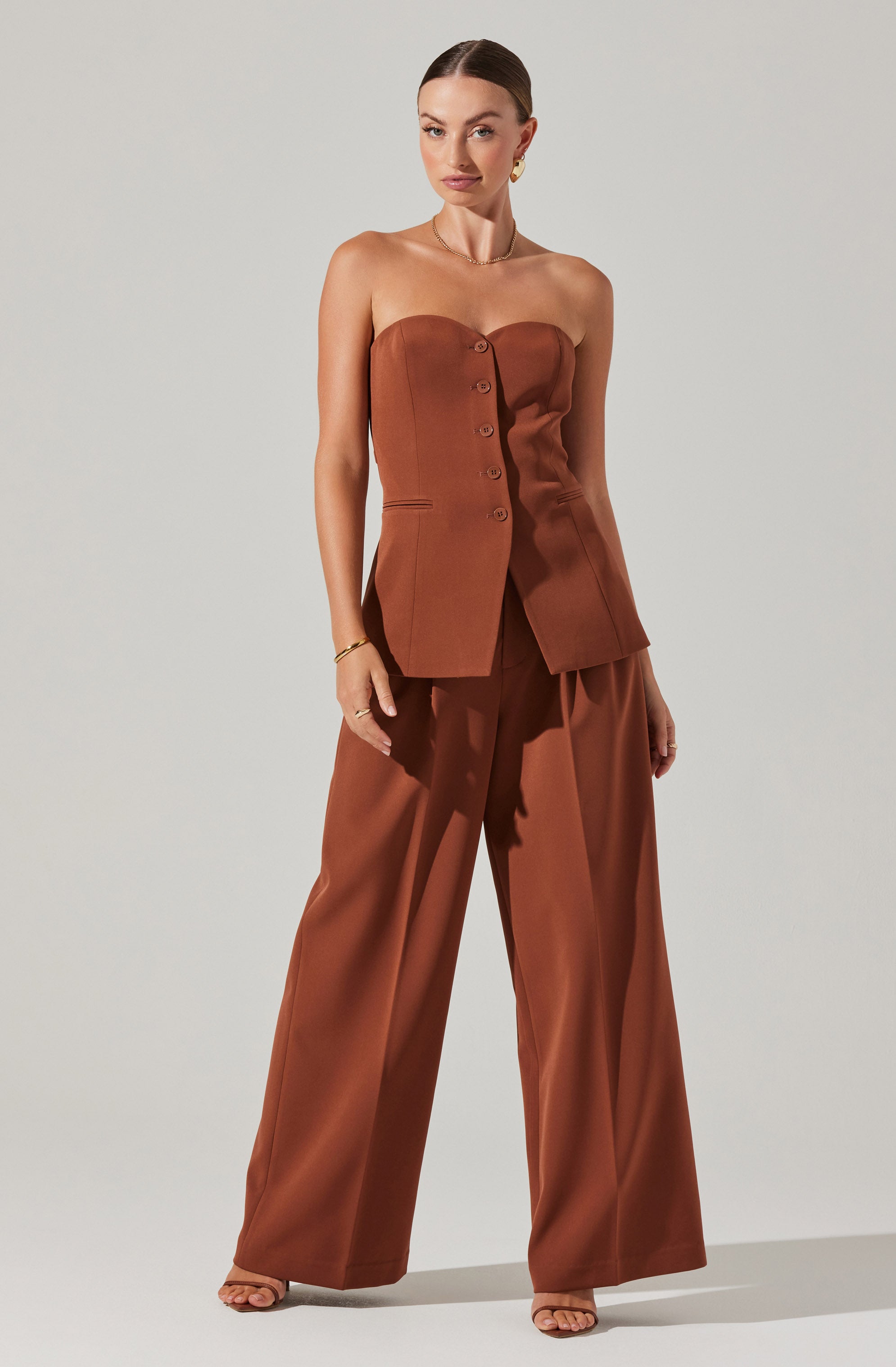 A woman is wearing a strapless, button-front, rust-colored tailored vest with matching wide-leg wide-leg trousers. The vest has a fitted silhouette with a slight peplum shape at the hips. The trousers are high-waisted and flow loosely. The outfit appears elegant and sophisticated.