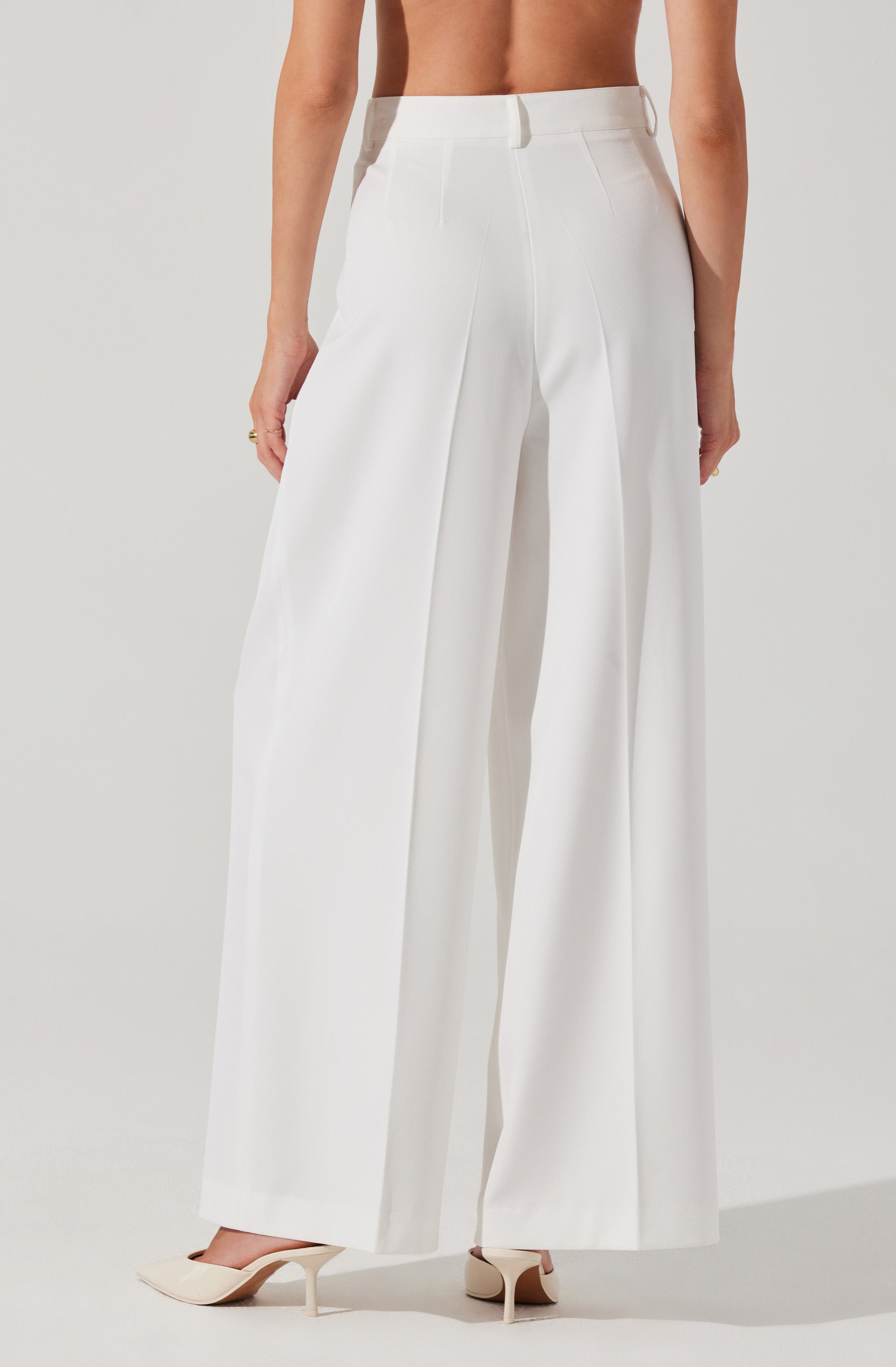 A person is wearing high-waisted, wide-leg white trousers made of a smooth, crisp fabric. The trousers feature a flat front with a waistband that has belt loops, and they are tailored with pleats running down the front of each leg. The person is also wearing beige pointed-toe high heels.