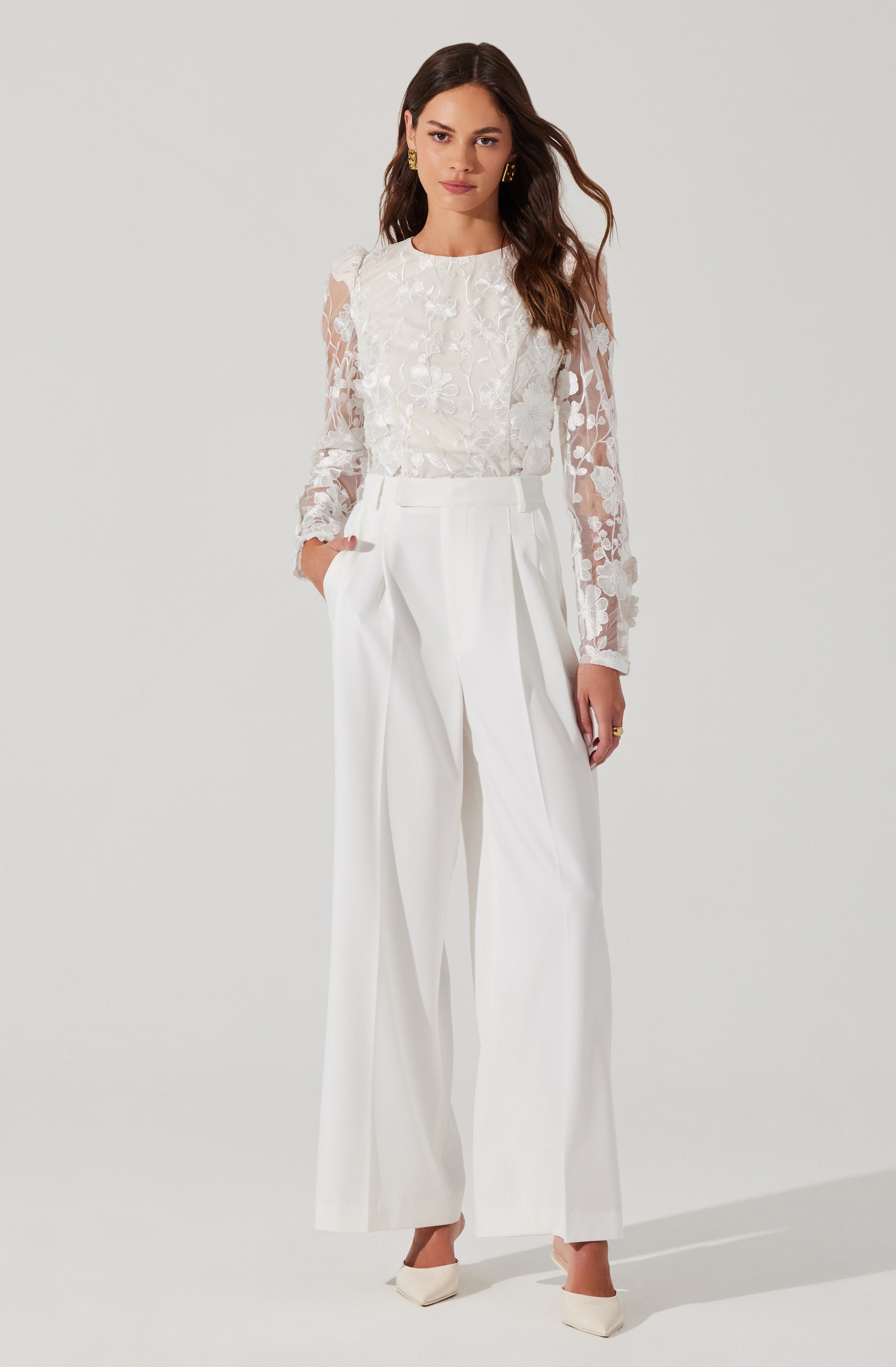 A woman is wearing a white long-sleeved blouse with lace floral patterns on the sleeves and front. She pairs it with high-waisted, wide-leg white trousers that feature pleats at the waist. She completes the outfit with white heels.