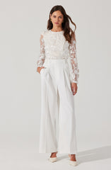 A woman is wearing a white long-sleeved blouse with lace floral patterns on the sleeves and front. She pairs it with high-waisted, wide-leg white trousers that feature pleats at the waist. She completes the outfit with white heels.