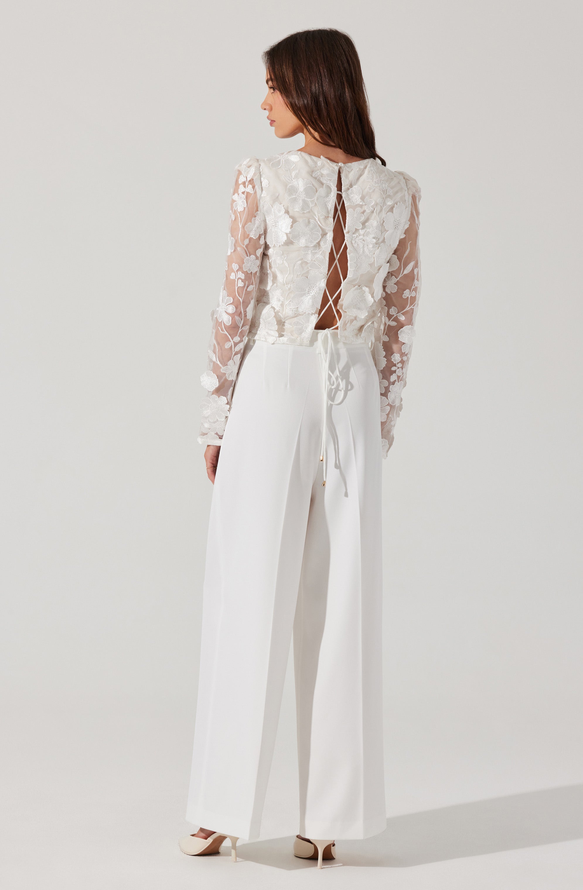 A woman is wearing a long-sleeved, sheer lace top with floral embroidery, featuring a cutout back with a crisscross lace-up detail. She pairs it with high-waisted, wide-leg white trousers and beige heels.
