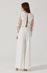 A woman is wearing a long-sleeved, sheer lace top with floral embroidery, featuring a cutout back with a crisscross lace-up detail. She pairs it with high-waisted, wide-leg white trousers and beige heels.