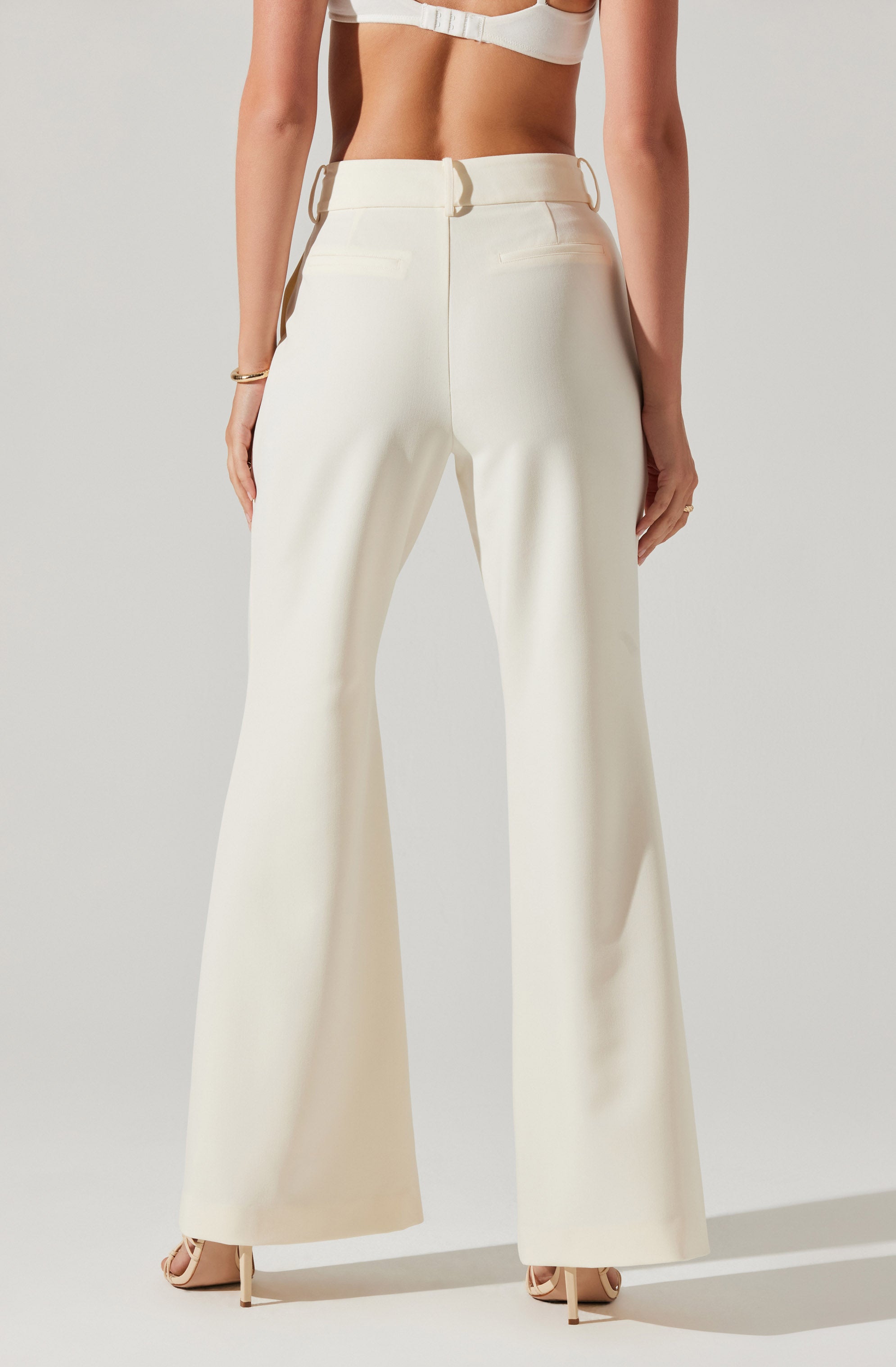 The image shows a person wearing high-waisted, wide-leg cream-colored trousers with belt loops, positioned against a plain light background. The person is also wearing a matching cream-colored cropped top and nude-colored heels.