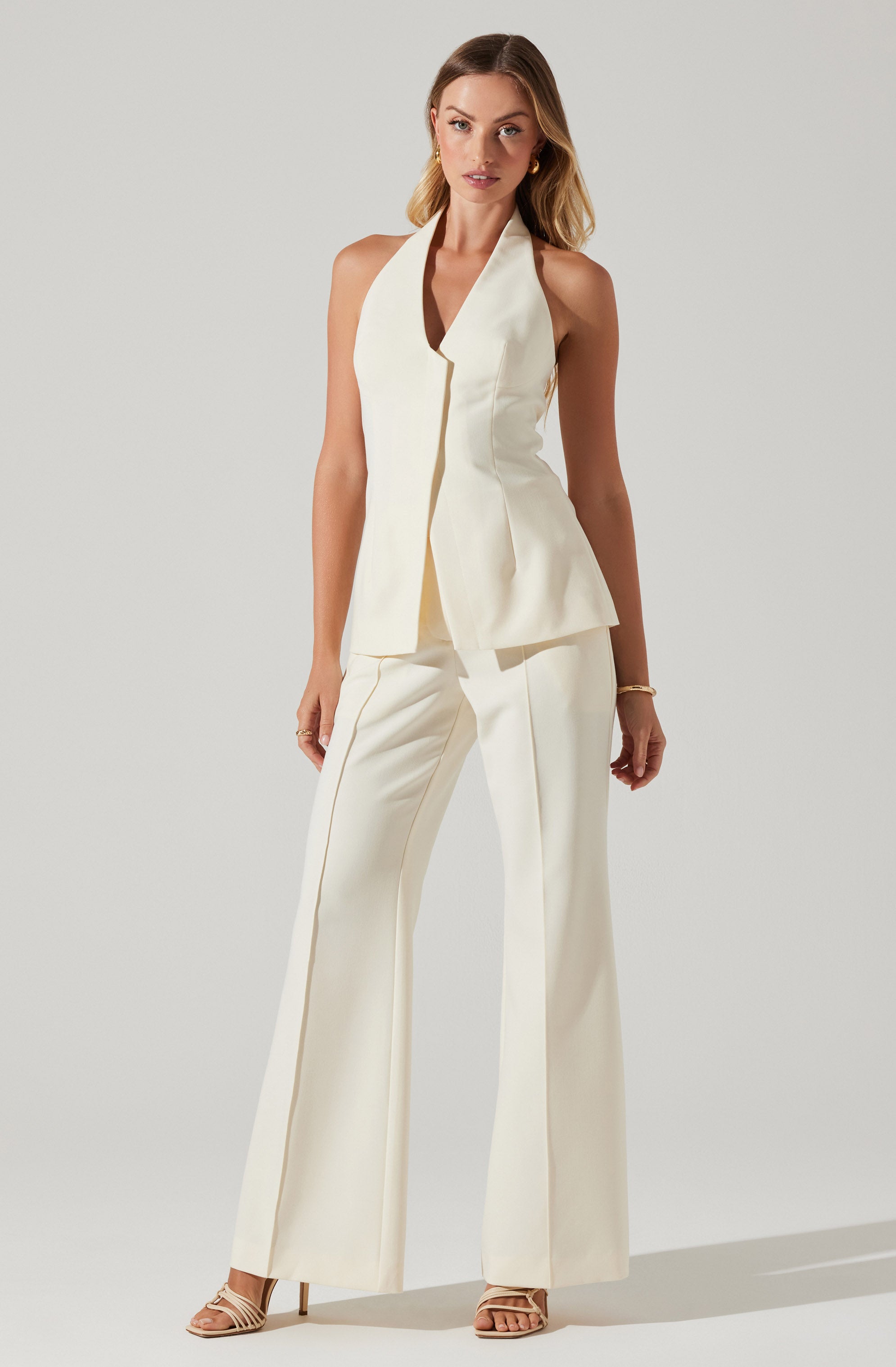 A woman is wearing an elegant, sleeveless cream-colored pantsuit. The top features a high collar and a deep V neckline, with a tailored, structured fit that flares slightly at the hem. The matching wide-leg trousers have a relaxed fit and a subtle crease down the front. She is also wearing nude strappy high-heeled sandals and simple gold jewelry.