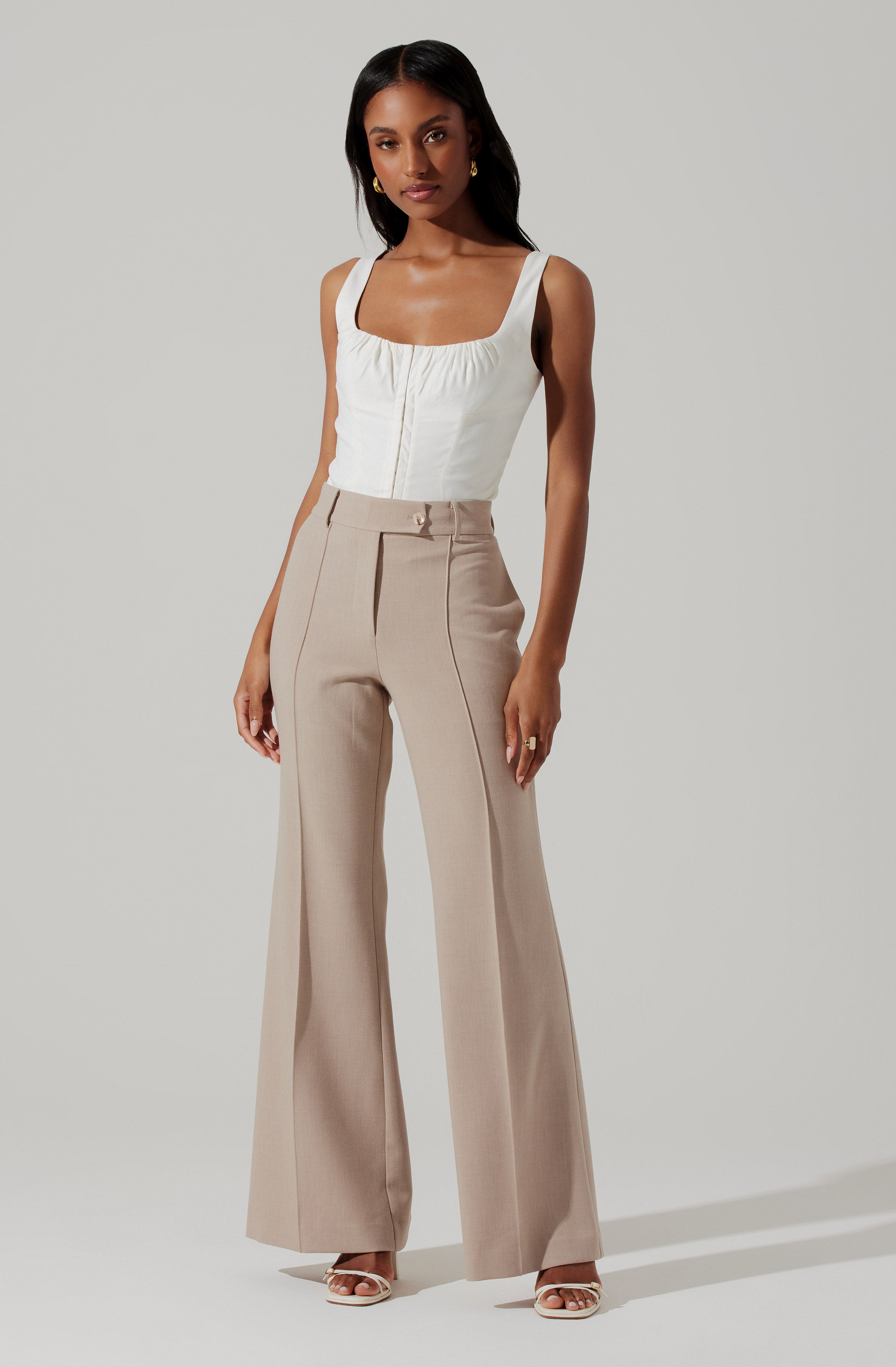 A woman is wearing a sleeveless, cream-colored top with a square neckline and a front zipper, paired with high-waisted, wide-leg beige trousers with vertical seam details. She is also wearing open-toe high-heeled shoes and gold hoop earrings.