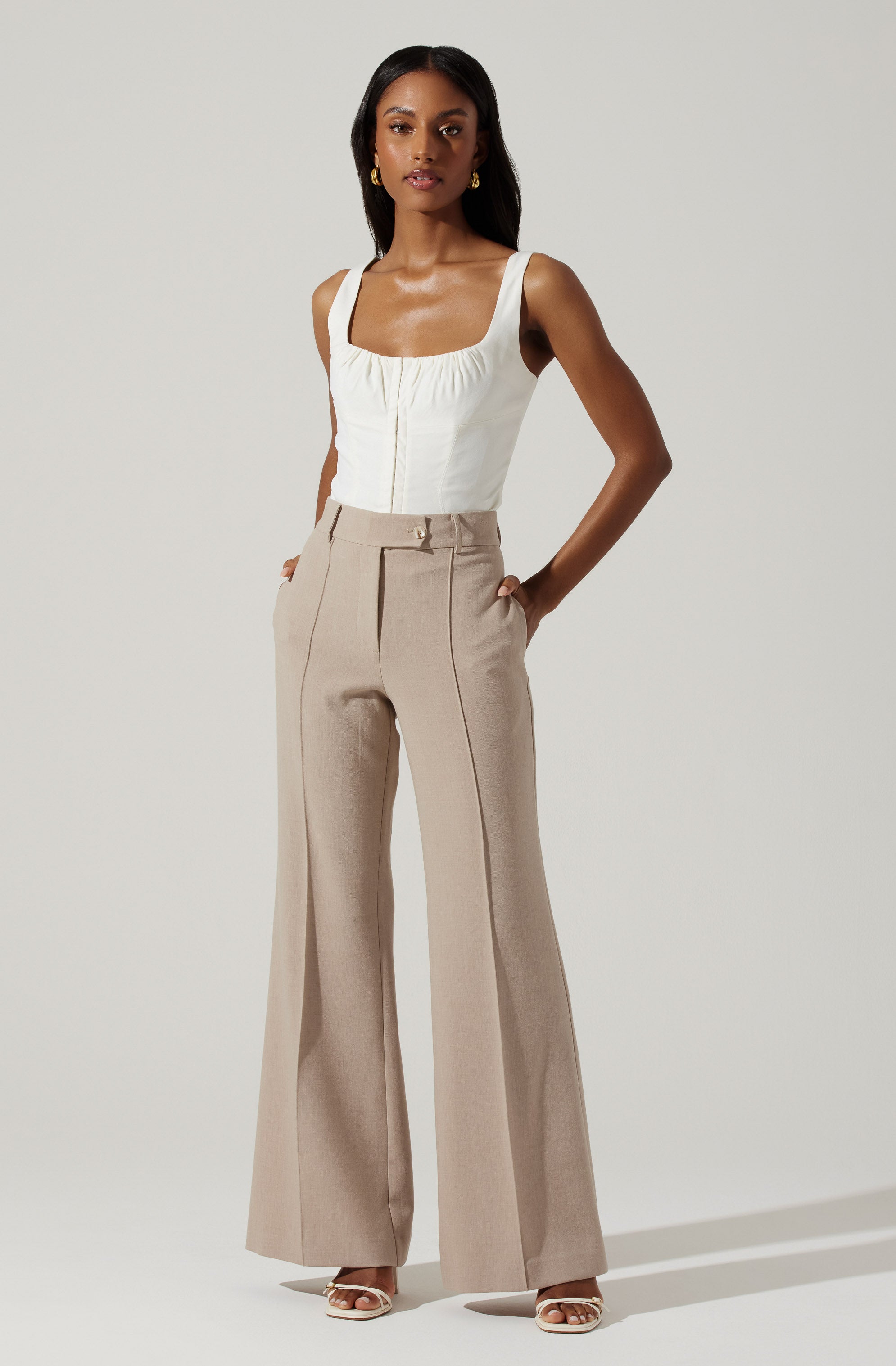 A woman is wearing a fitted white sleeveless top with wide straps and a gathered detail at the front, paired with high-waisted beige wide-leg trousers that have a classic waistband, front pleats, and a hidden front zipper. She completes the outfit with gold earrings and white open-toe heels.