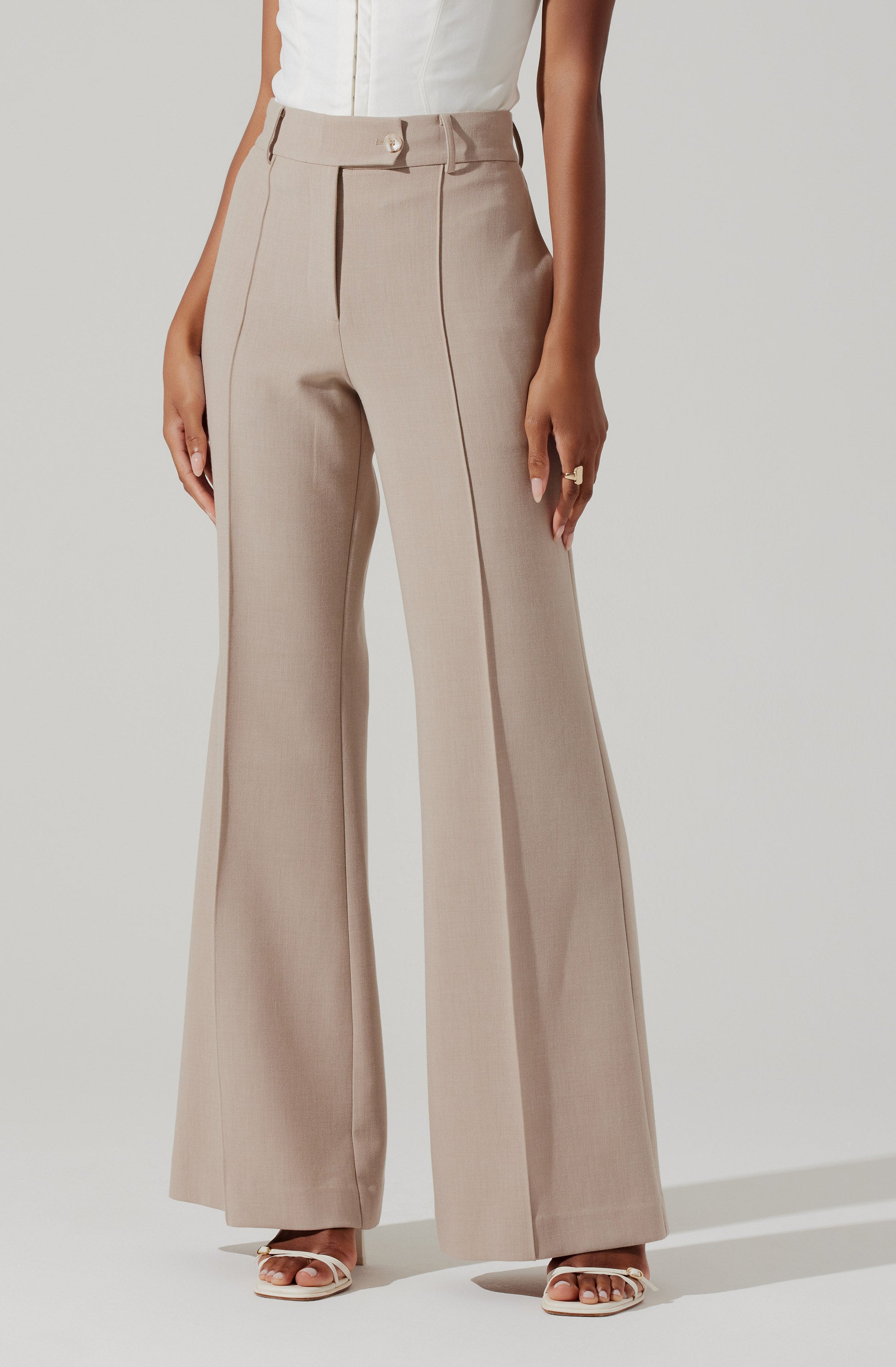 A person is wearing high-waisted beige wide-leg trousers with a front zipper and hook closure, along with a sleeveless white top. The trousers have a tailored fit with vertical seams and a subtle plaid pattern. The person is also wearing white open-toe heels.