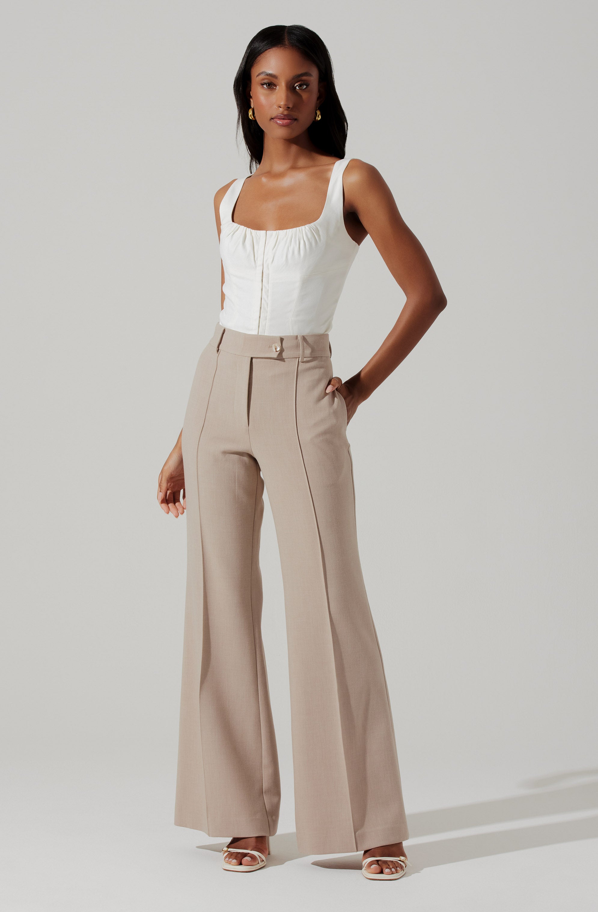A woman is wearing a sleeveless, white top with a square neckline and gathered front detail. She pairs it with high-waisted, wide-leg beige trousers featuring visible front pleats and vertical seams down the front of each leg. She completes her outfit with open-toe heels.