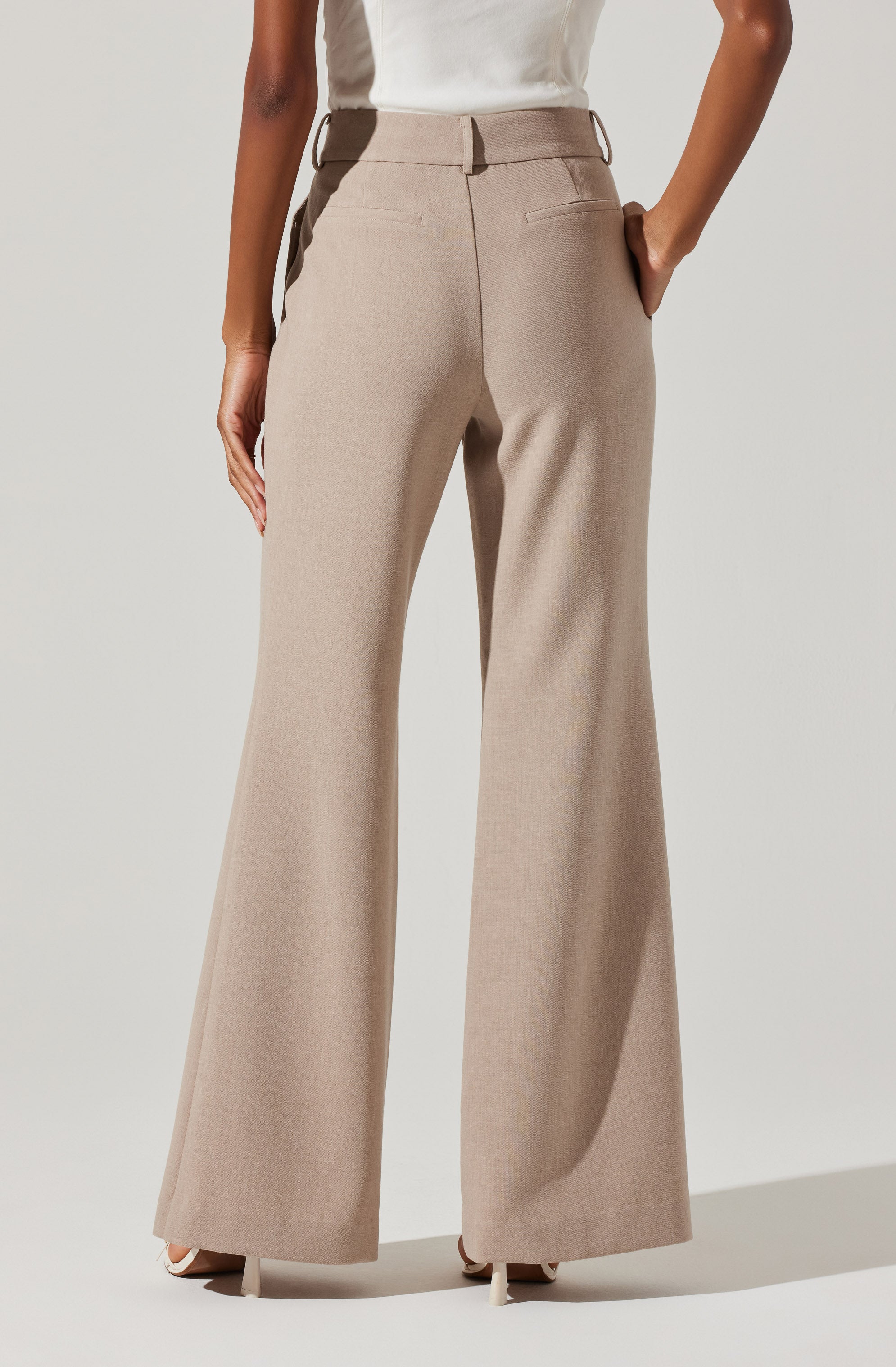 A person is wearing high-waisted, wide-leg beige dress pants with a subtle checkered pattern. The pants feature belt loops and back welt pockets. They are paired with a sleeveless white top tucked into the pants. The person has one hand in their pocket and the other hanging naturally by their side.