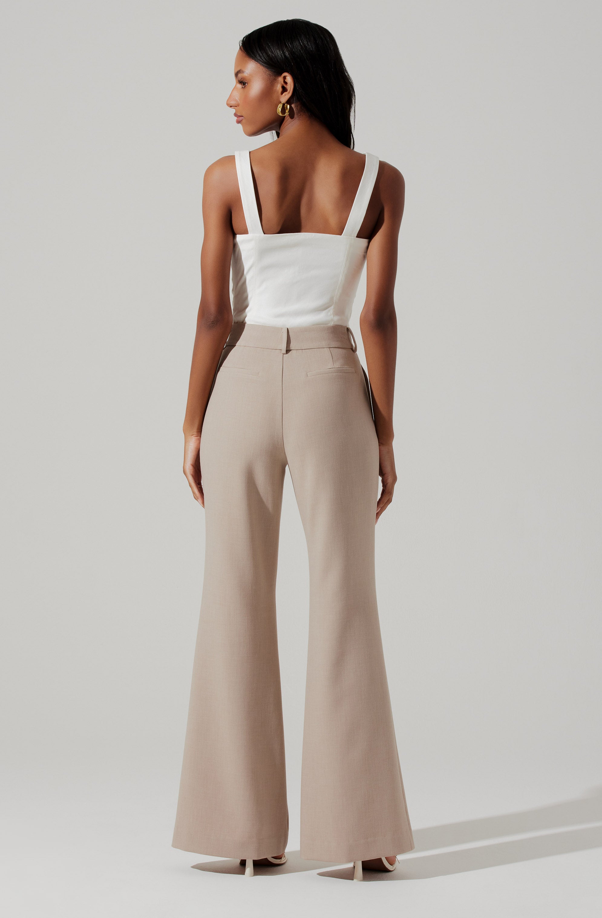 A woman is wearing a white sleeveless top with wide shoulder straps and a square neckline, paired with high-waisted, wide-leg beige pants.