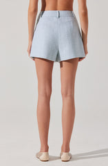 Light blue textured shorts with a high waist, front pleats, and belt loops. The shorts have a tailored fit with a slightly flared leg opening. The model is also wearing beige shoes with small decorative elements.