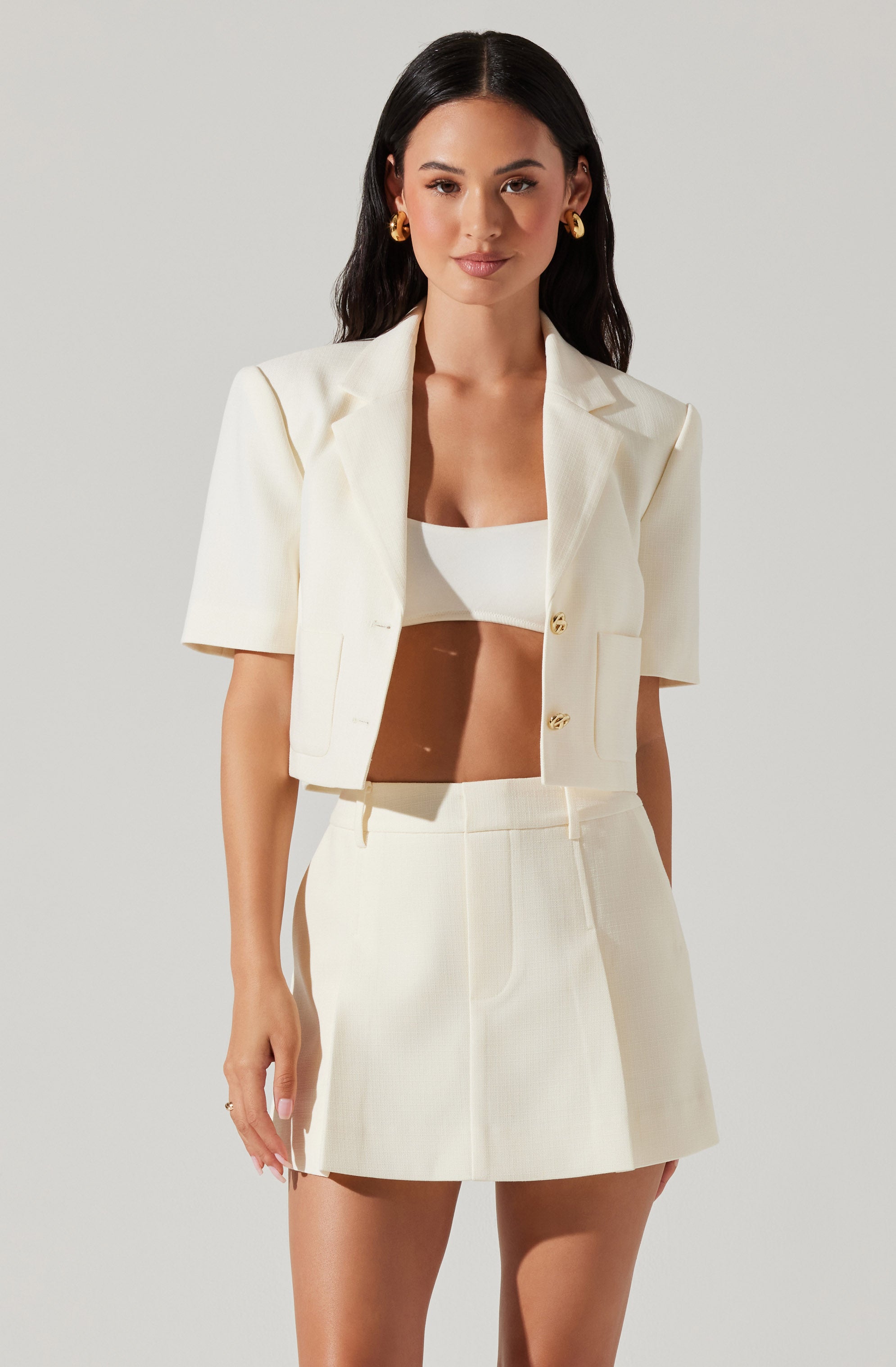 A woman is wearing a cream-colored, short-sleeved blazer with padded shoulders and gold buttons. She has paired it with a matching cream-colored mini skirt and a white crop top underneath. She accessorizes with large gold hoop earrings.