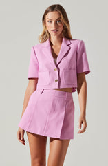 A woman is wearing a light pink cropped blazer with short sleeves and a notched lapel collar, featuring two large front pockets and two decorative buttons. She pairs it with a matching high-waisted pink mini skirt with vertical seam details.