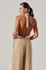A woman is wearing a sleeveless, backless white halter top with a tied bow at the neck. She pairs it with high-waisted, beige wide-leg trousers. She accessorizes with large gold hoop earrings.