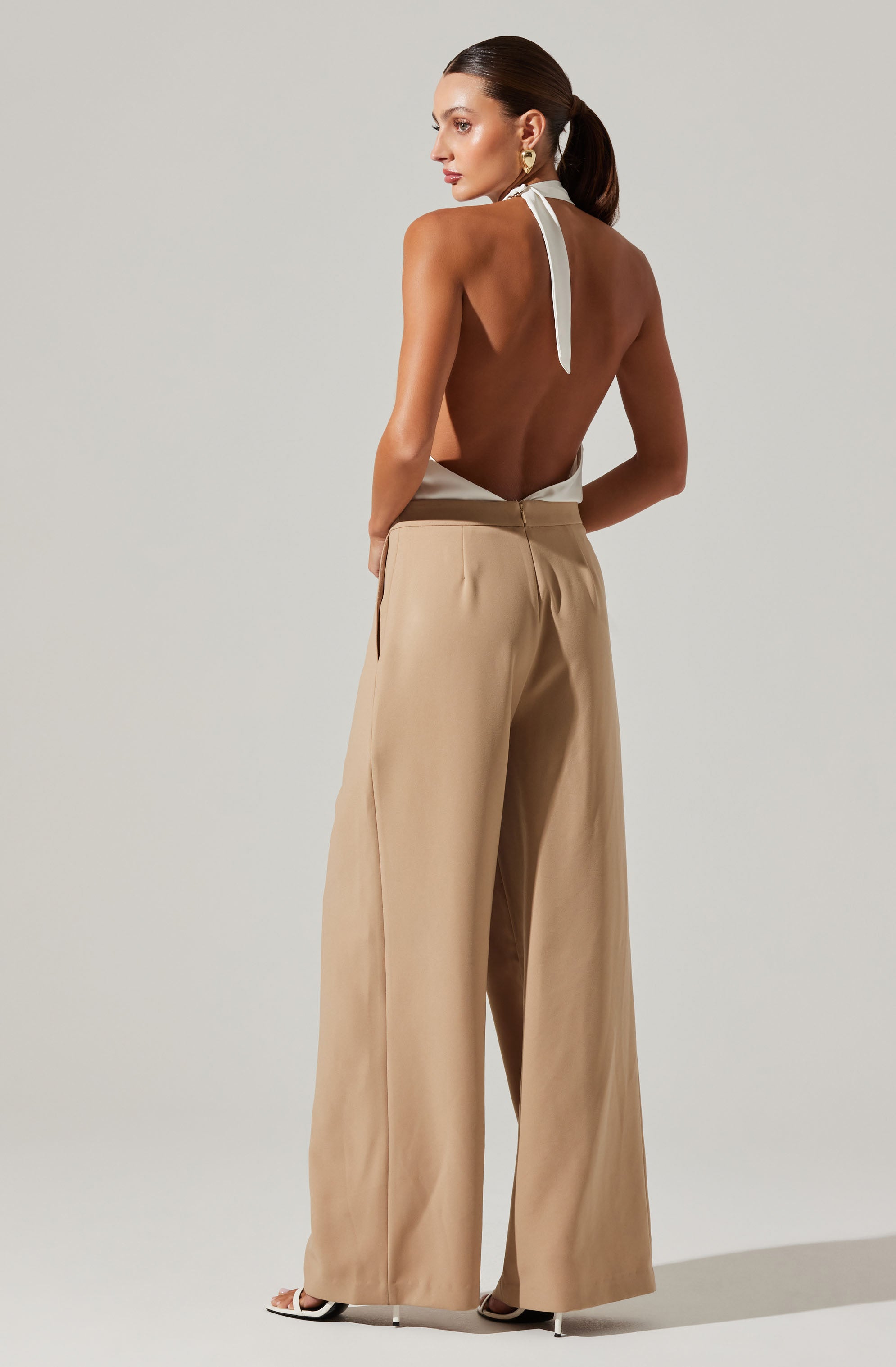 A woman is wearing high-waisted, wide-leg beige trousers paired with a sleeveless, backless top that ties around the neck with a white strap. She is accessorized with large gold earrings and stands against a plain, light-colored background.