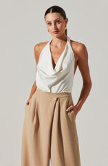 A woman is wearing a sleeveless white top with a draped neckline and beige high-waisted wide-leg trousers with pleats and a button closure.