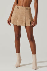A woman is wearing a beige, textured, high-waisted mini skirt with decorative buttons at the waist and front pleats. She is also wearing light-colored ankle boots with a shiny finish.