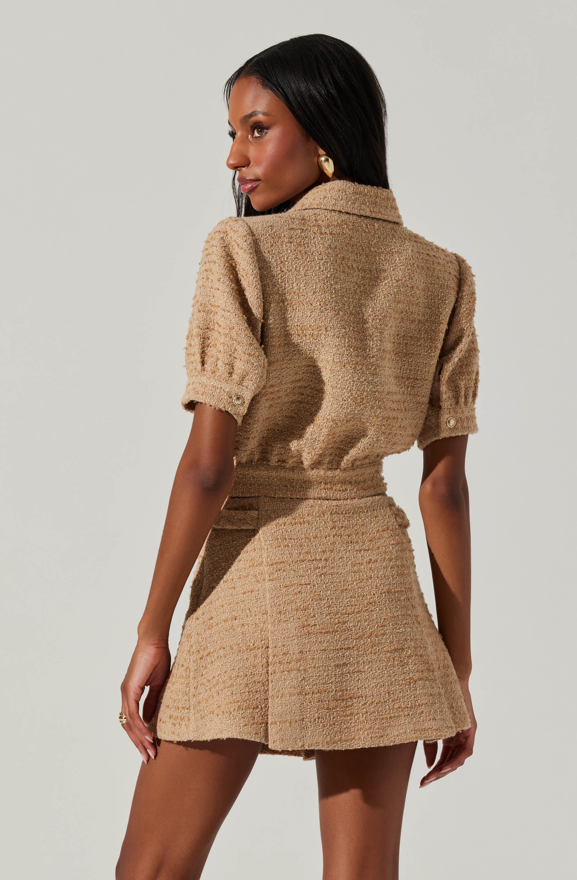 A woman is wearing a matching beige textured blazer and skirt set. The blazer has short puffed sleeves with buttoned cuffs and a collar, while the skirt is high-waisted and slightly flared. She has long, straight black hair and is accessorized with large, gold hoop earrings.