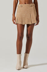 A woman is wearing a beige textured mini skirt with a high waist, featuring two decorative buttons on the waistband and two front pleats. She is also wearing knee-high, cream-colored boots with a zipper on the side.
