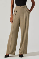 A person is wearing high-waisted, wide-leg beige trousers with pleats at the front, paired with a black top. The trousers feature belt loops and a front button closure. The individual is standing with one hand in the pocket of the trousers and the other hand resting by their side. They are also wearing black pointed-toe heels.