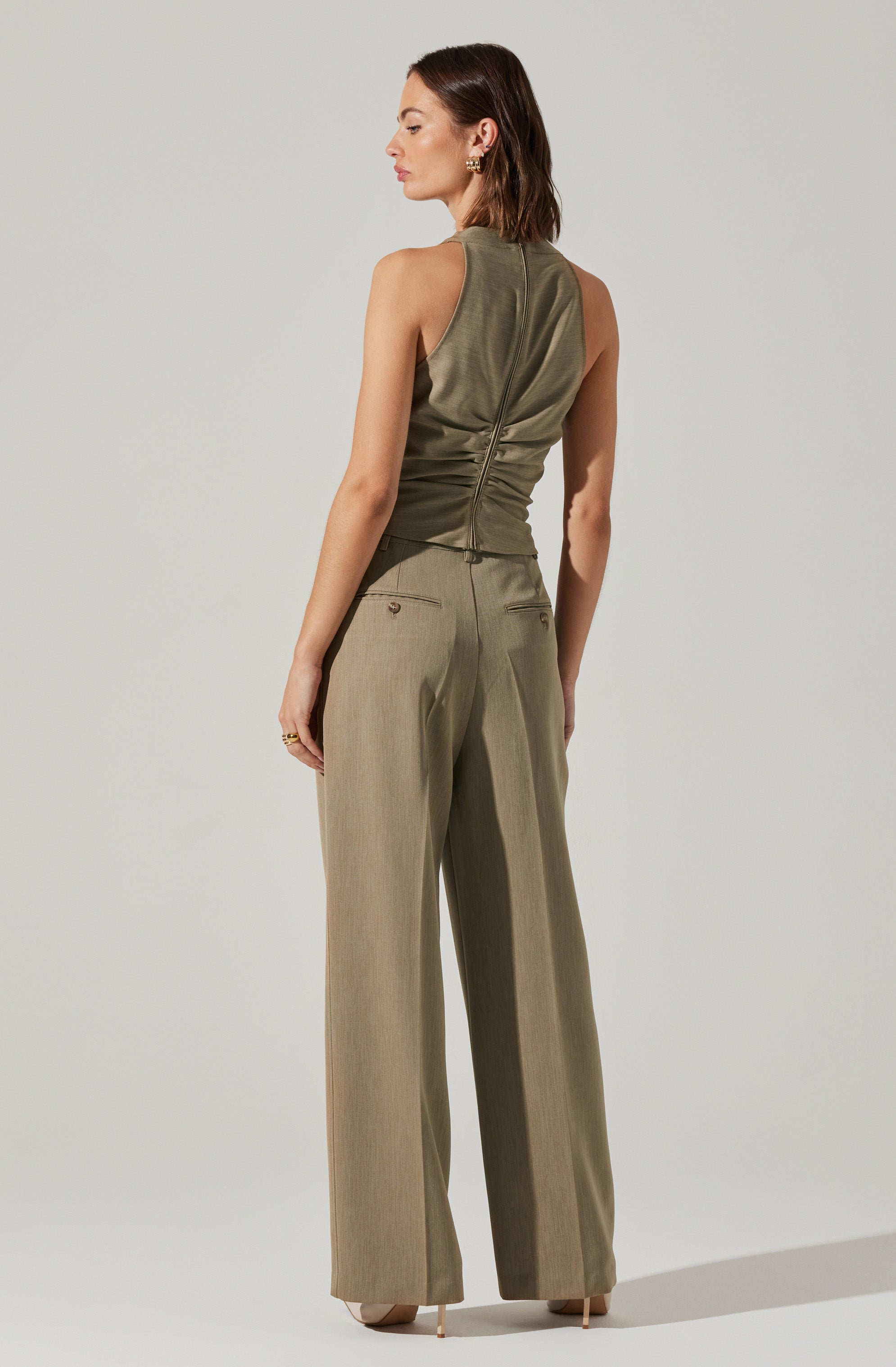 A woman is wearing a sleeveless, olive-green top with a high neckline and a central zipper at the back. She is also wearing high-waisted, wide-leg beige trousers with back welt pockets.