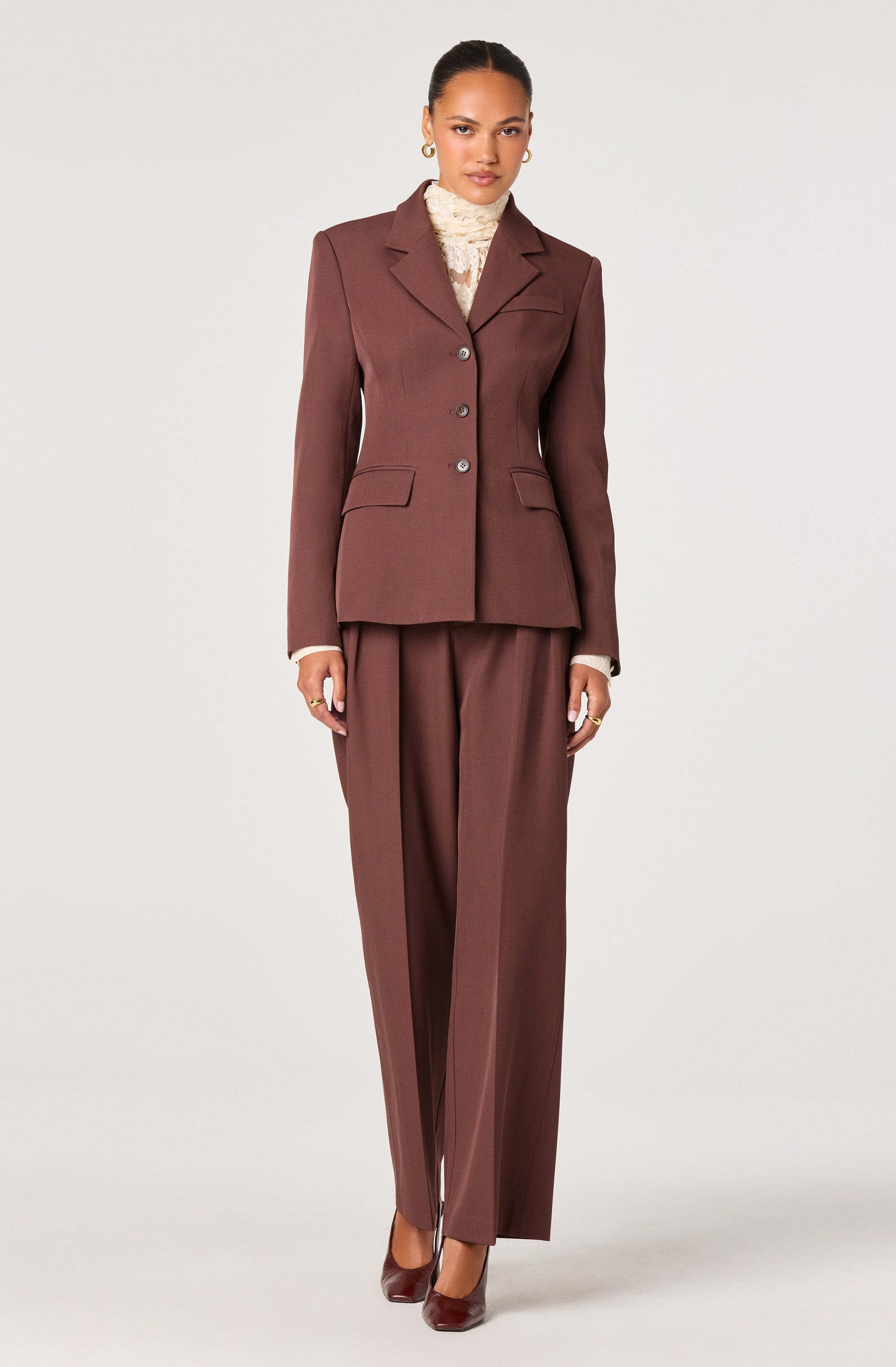 A woman is wearing a fitted, brown tailored blazer with notch lapels, three buttons down the front, and two flap pockets at the waist. She has on matching wide-leg brown trousers with a high waist and pleats. Under the blazer, she is wearing a cream-colored, textured turtleneck sweater. She completes the look with dark brown pointed-toe heels and gold earrings.