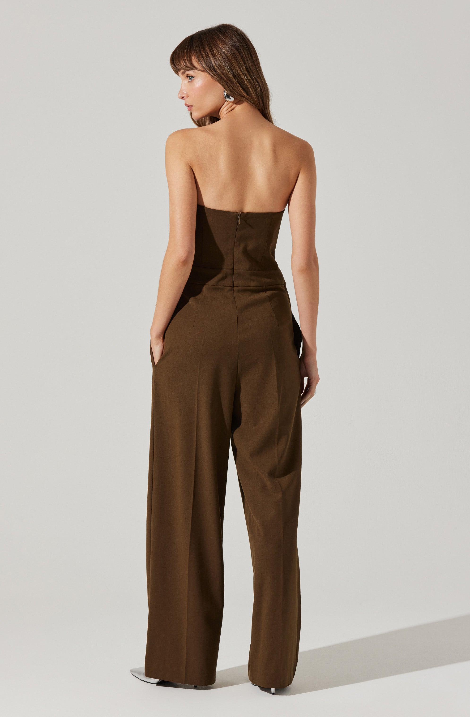 A woman is wearing a strapless, brown jumpsuit with wide-leg pants. The fabric appears smooth and slightly shiny. The back of the jumpsuit features a concealed zipper running down the center. The woman has her hands in her pockets and is standing against a plain, light gray background.
