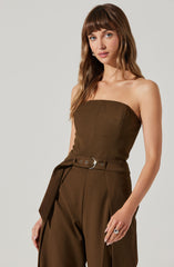 A woman is wearing a strapless, brown jumpsuit with a fitted bodice and high-waisted wide-leg pants. The jumpsuit features a matching belt with a silver buckle at the waist.
