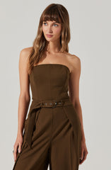 A woman is wearing a strapless brown jumpsuit with a fitted bodice and wide-leg pants. The jumpsuit features a belt at the waist with a silver buckle and decorative grommets, and the pants have pleats at the front.