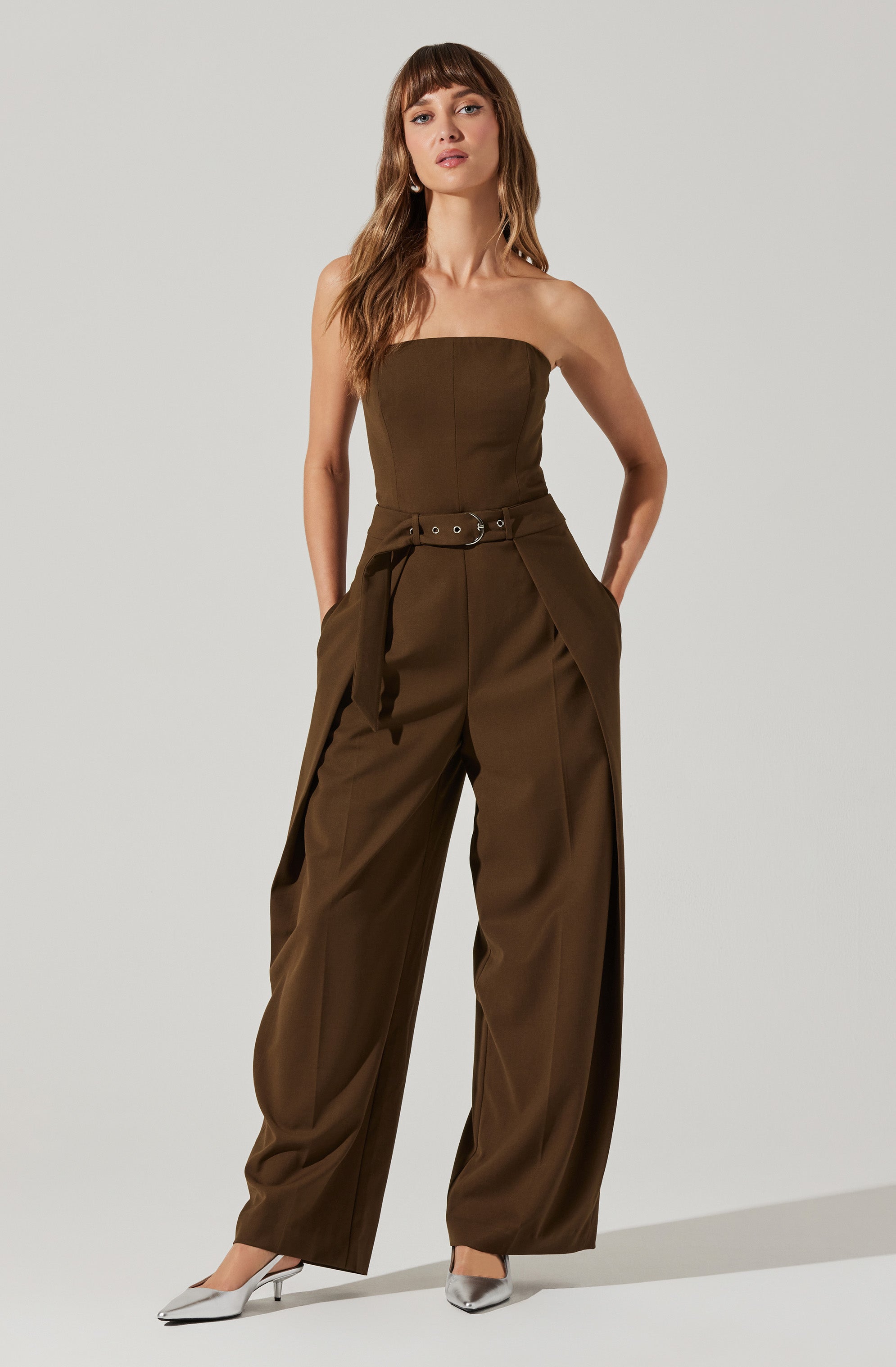 A woman is wearing an olive-green strapless jumpsuit with a fitted bodice and wide-leg pants. The jumpsuit features a matching fabric belt with grommet details at the waist, and the pants have pleats at the hips. She is also wearing pointed-toe silver heels.
