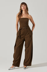 A woman is wearing an olive-green strapless jumpsuit with a fitted bodice and wide-leg pants. The jumpsuit features a matching fabric belt with grommet details at the waist, and the pants have pleats at the hips. She is also wearing pointed-toe silver heels.