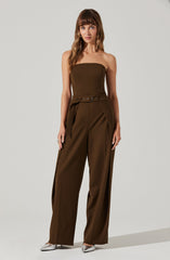 A woman is wearing a strapless, fitted brown jumpsuit with wide-leg pants. The jumpsuit features a belt at the waist with a metal buckle. She is also wearing pointed silver heels.