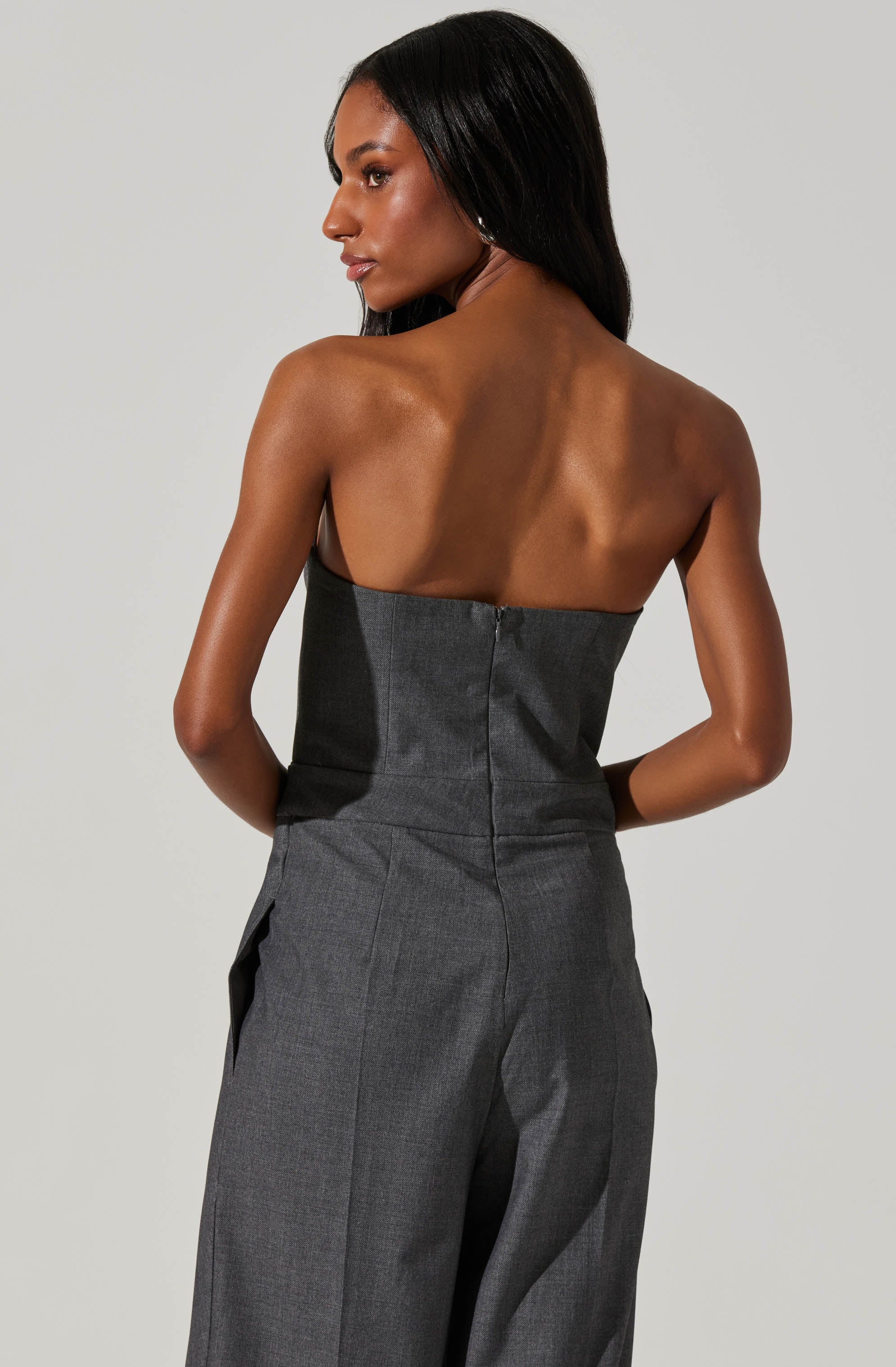 A woman with long black hair is wearing a strapless, dark gray jumpsuit. The jumpsuit is fitted at the waist with a visible zipper at the back and has wide-legged pants. Her skin is medium brown, and she is posed with her back slightly turned, looking to the side against a plain, light background.