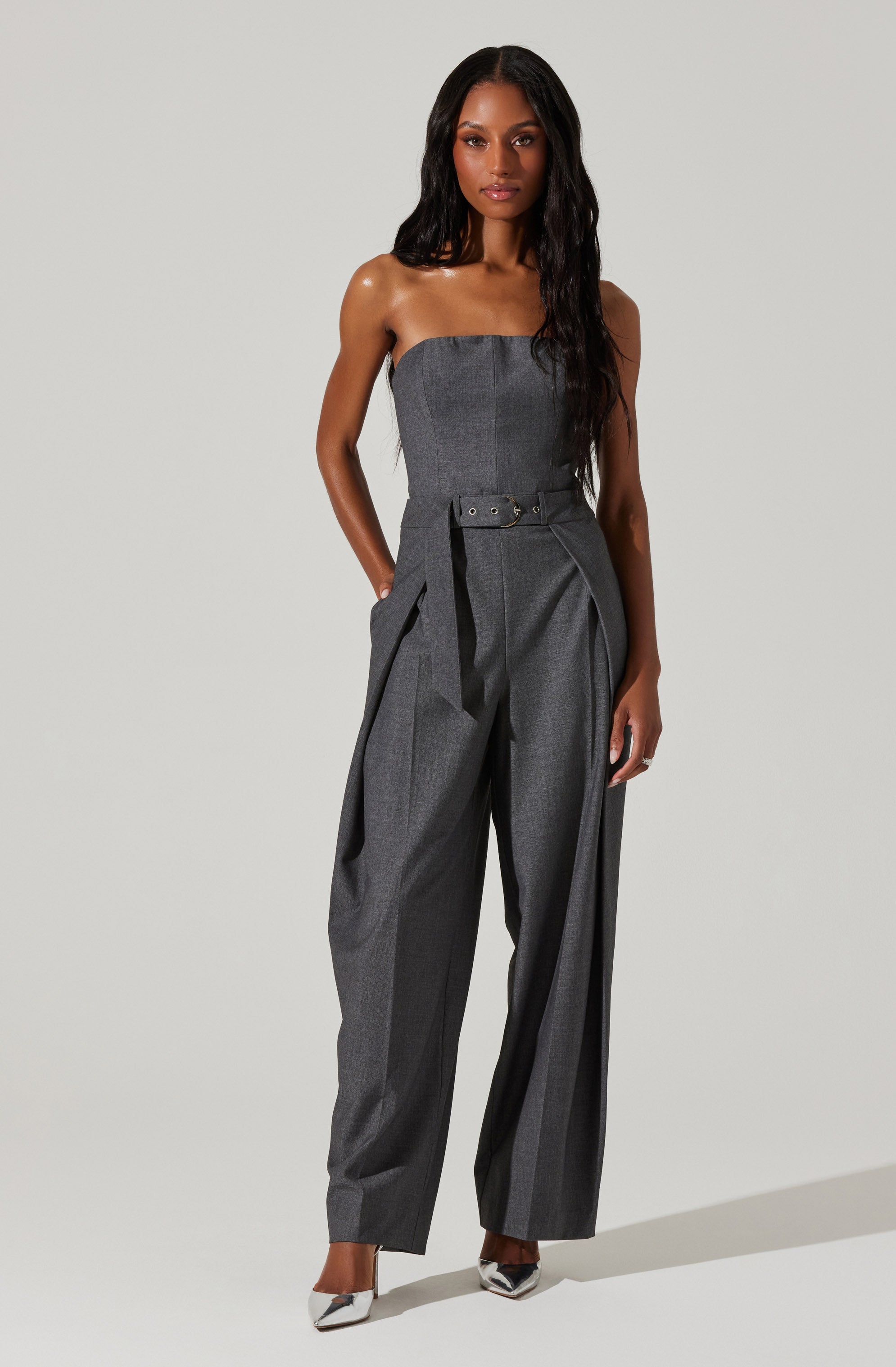 A woman is wearing a strapless, dark gray jumpsuit with wide-leg pants. The jumpsuit features a built-in belt at the waist with a rectangular buckle and pleated detailing on the pants. She has long, dark, wavy hair and is standing against a plain light background.