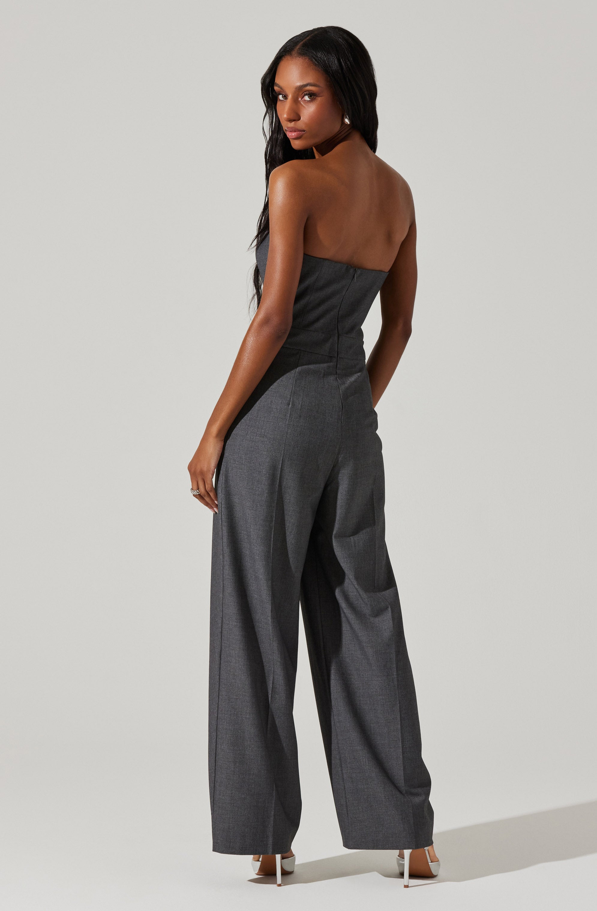 A woman is wearing a strapless jumpsuit with a fitted bodice. The top part of the jumpsuit is in a dark gray color, while the wide-legged pants are in a lighter gray shade. She is also wearing white high-heeled shoes.