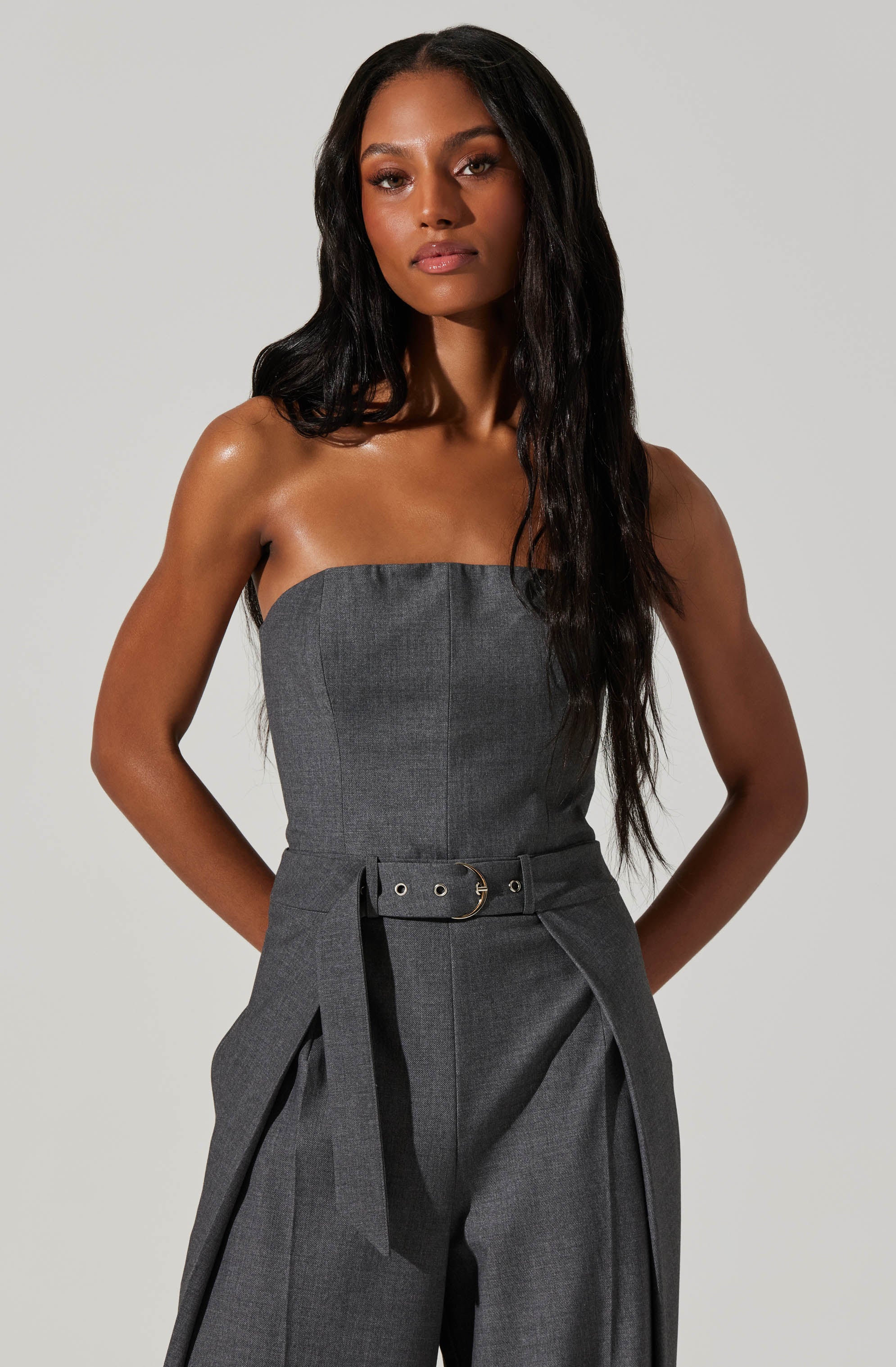A woman is wearing a strapless, fitted, gray jumpsuit with a wide belt that has grommets and a D-ring buckle. The jumpsuit features tailored, wide-legged trousers with pleats.