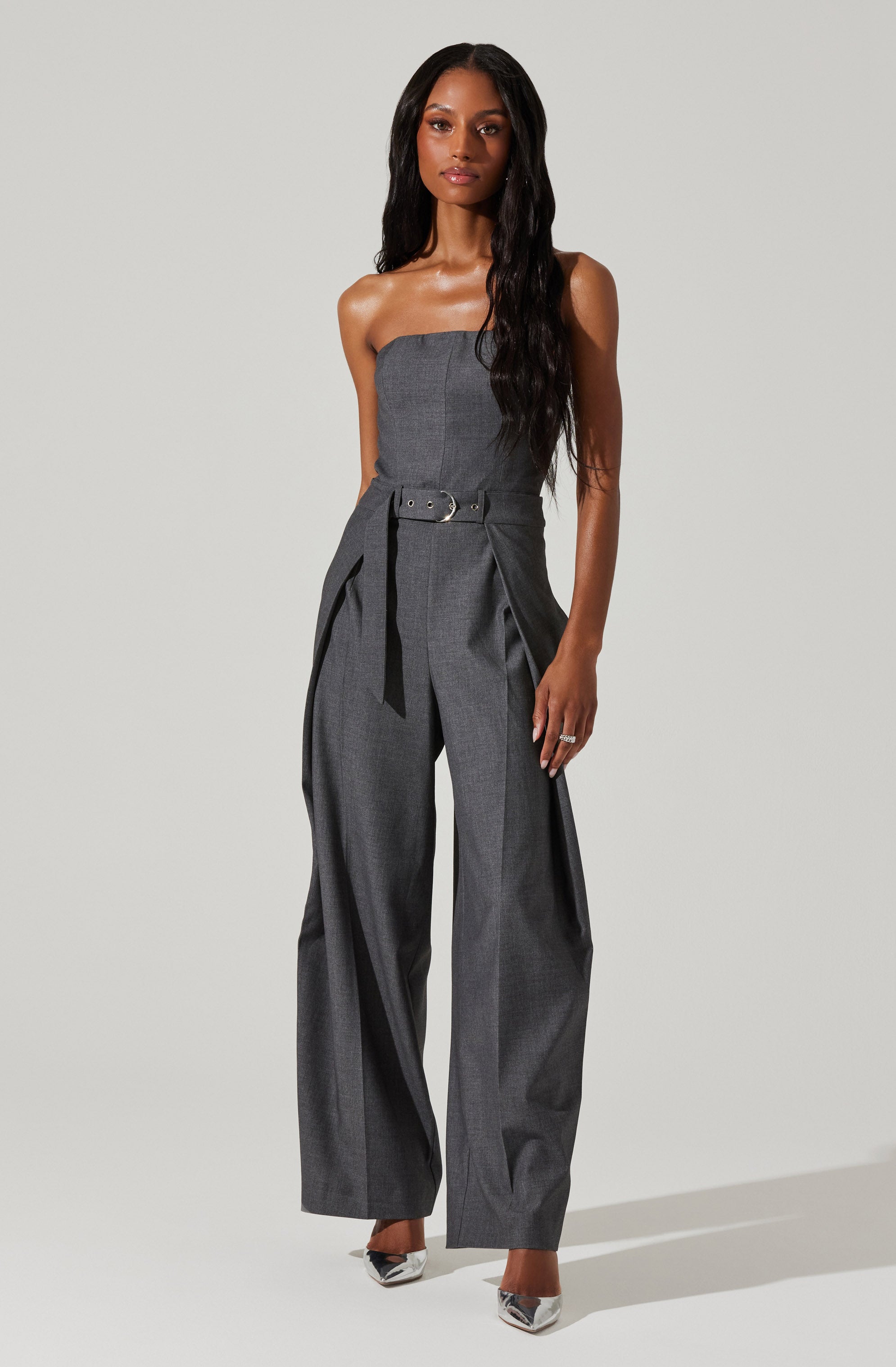 A woman is wearing a strapless, floor-length, high-waisted gray jumpsuit with wide-leg pants. The jumpsuit features a fitted bodice and a belt at the waist with a silver buckle. The fabric appears to be a classic suiting material. She is also wearing pointed-toe high heels with a metallic finish.
