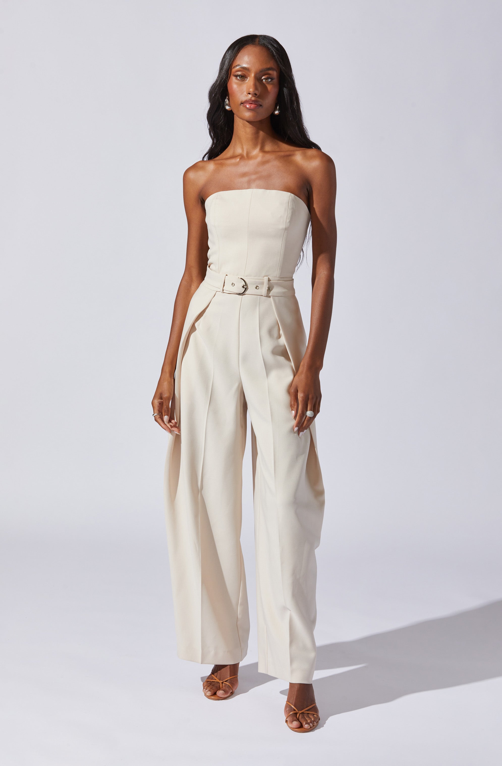 A woman is wearing a strapless, cream-colored jumpsuit with a fitted bodice and wide-leg pants. The jumpsuit features a matching belt with a circular buckle at the waist and pleats along the front of the pants. She is also wearing open-toe high-heeled sandals.