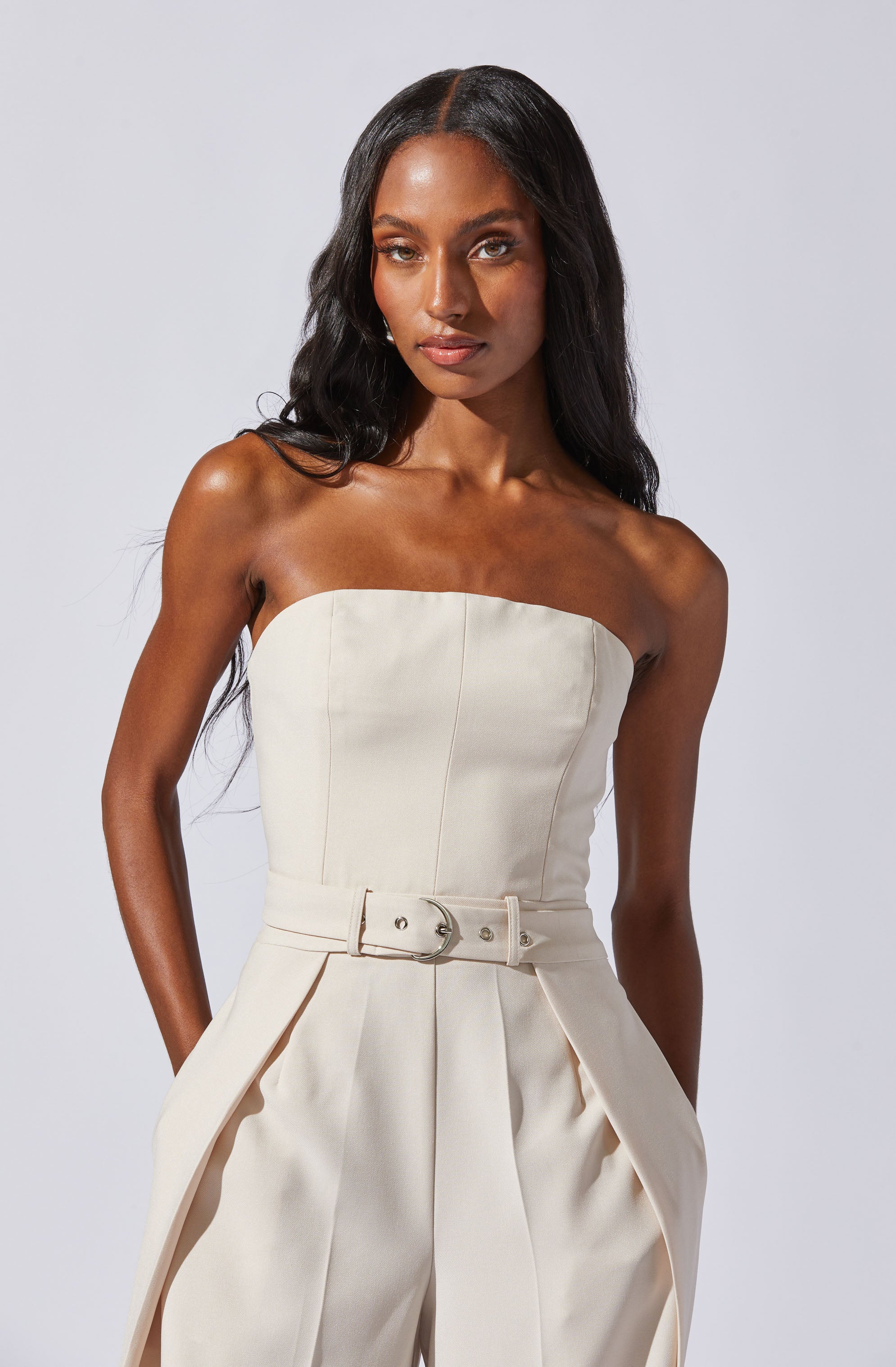 A woman wearing a strapless, cream-colored jumpsuit with a fitted bodice, high waist, and wide-leg pants featuring pleats. The jumpsuit has a matching belt with a silver buckle at the waist.